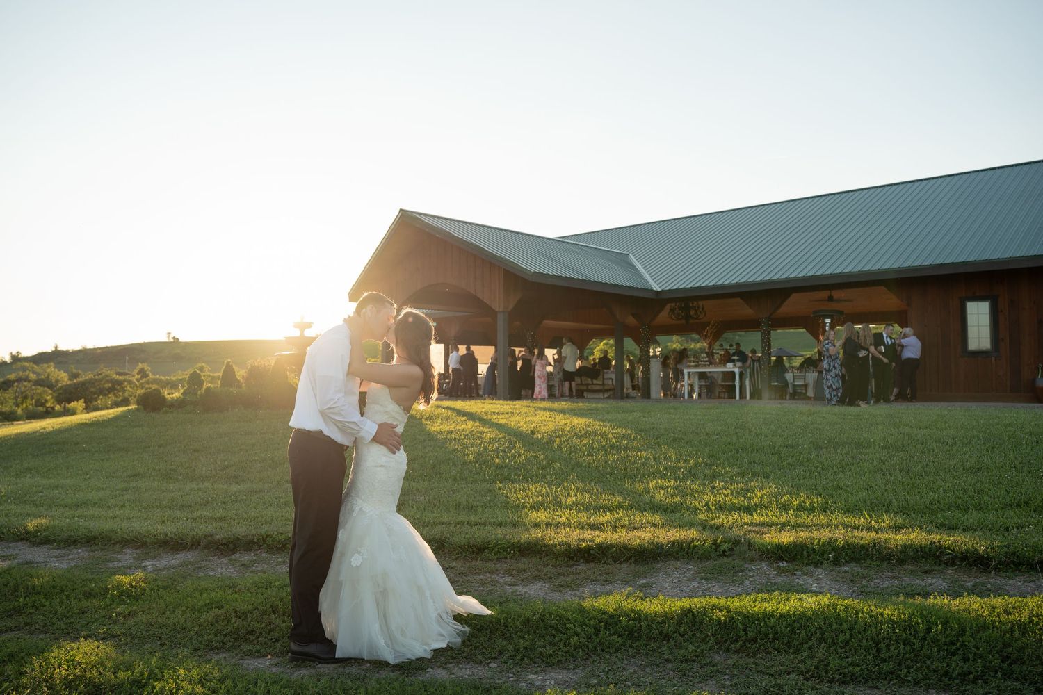 Bride and groom share romantic sunset kiss on green lawn outside rustic wedding venue.