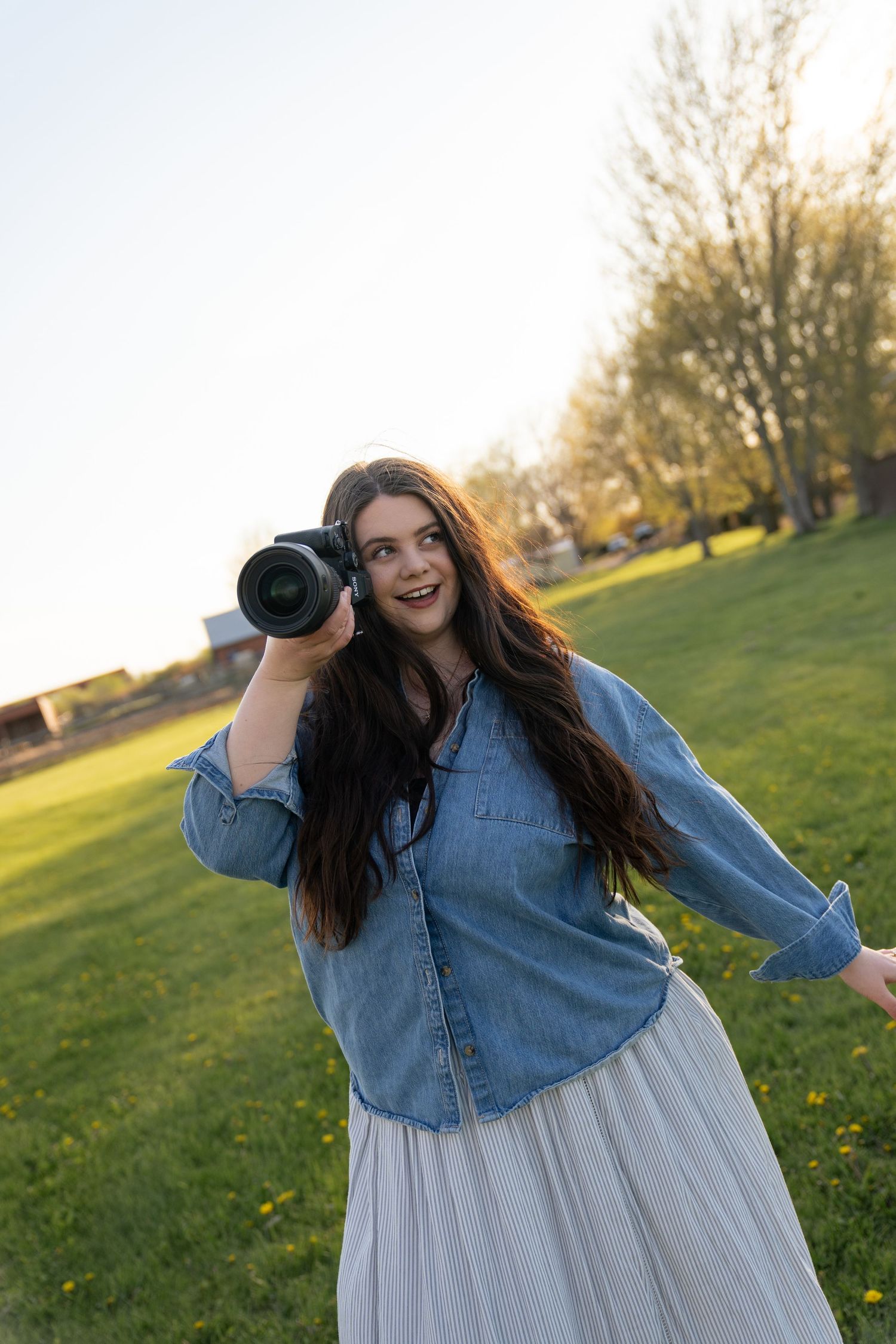 Smiling female photographer in denim shirt and white skirt holding camera in a sunny green field.