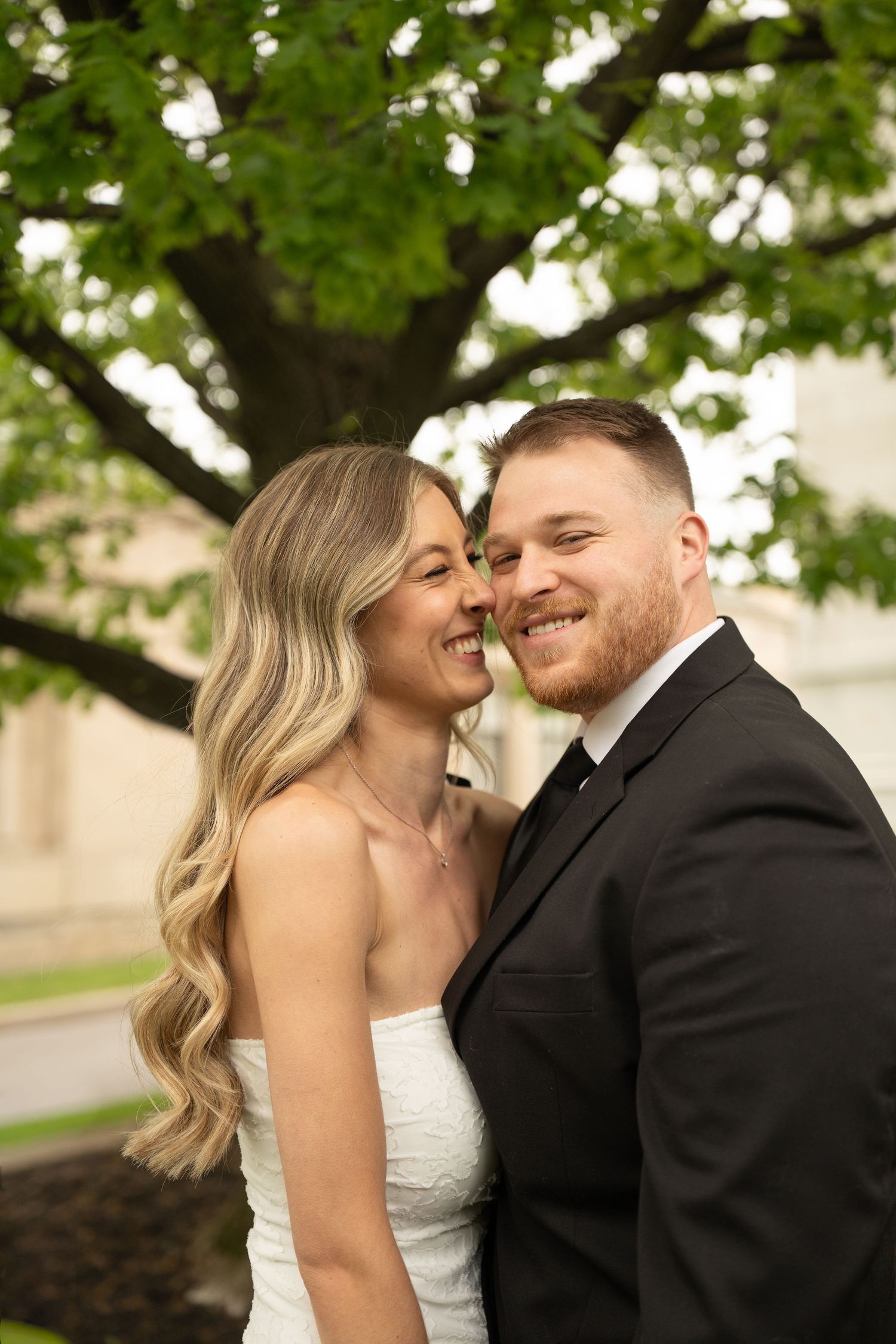 Smiling wedding couple embracing outdoors under a green tree, bride in strapless gown and groom in black suit.
