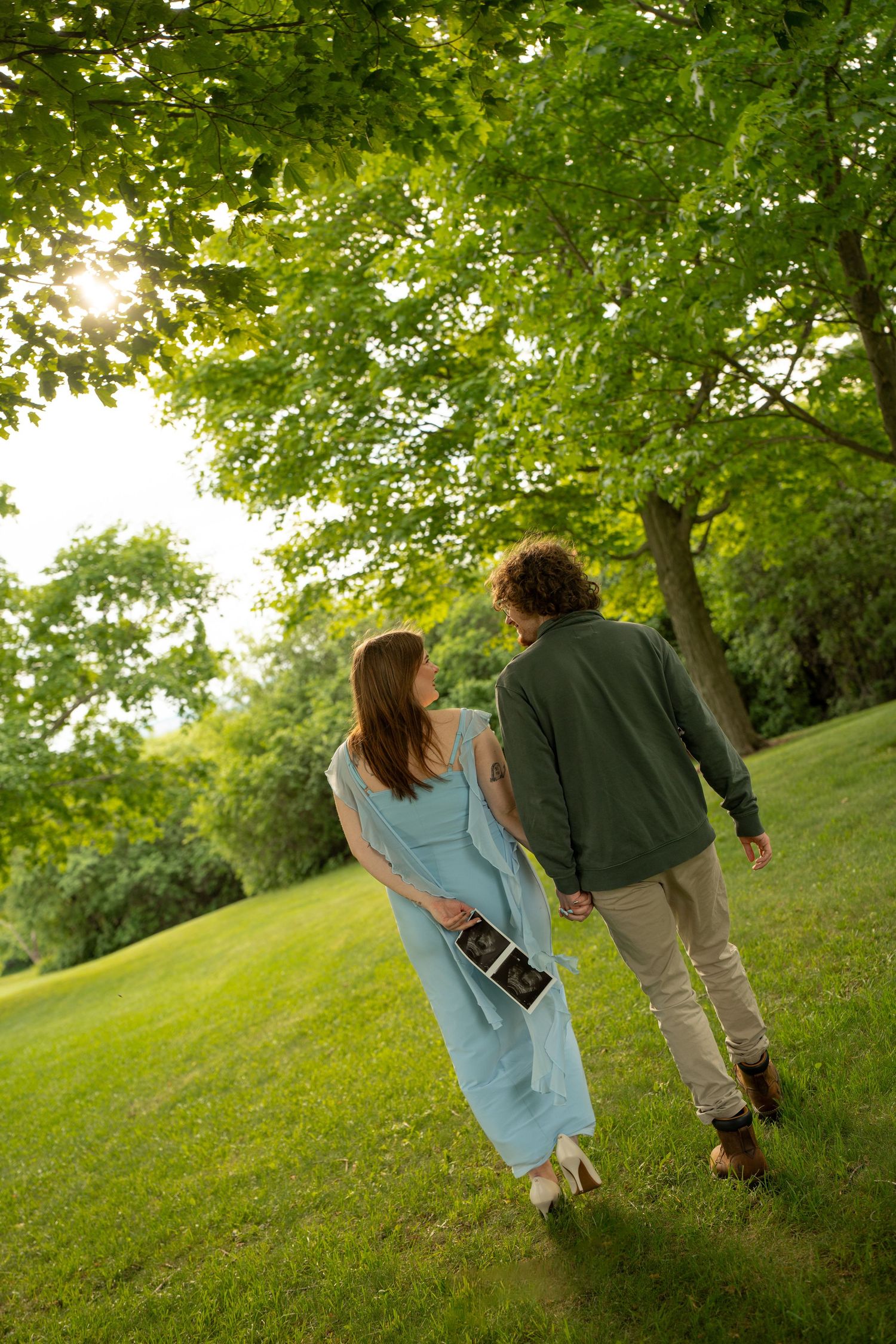 Pregnant couple holding ultrasound photo walking hand in hand through sunlit green park under large tree.