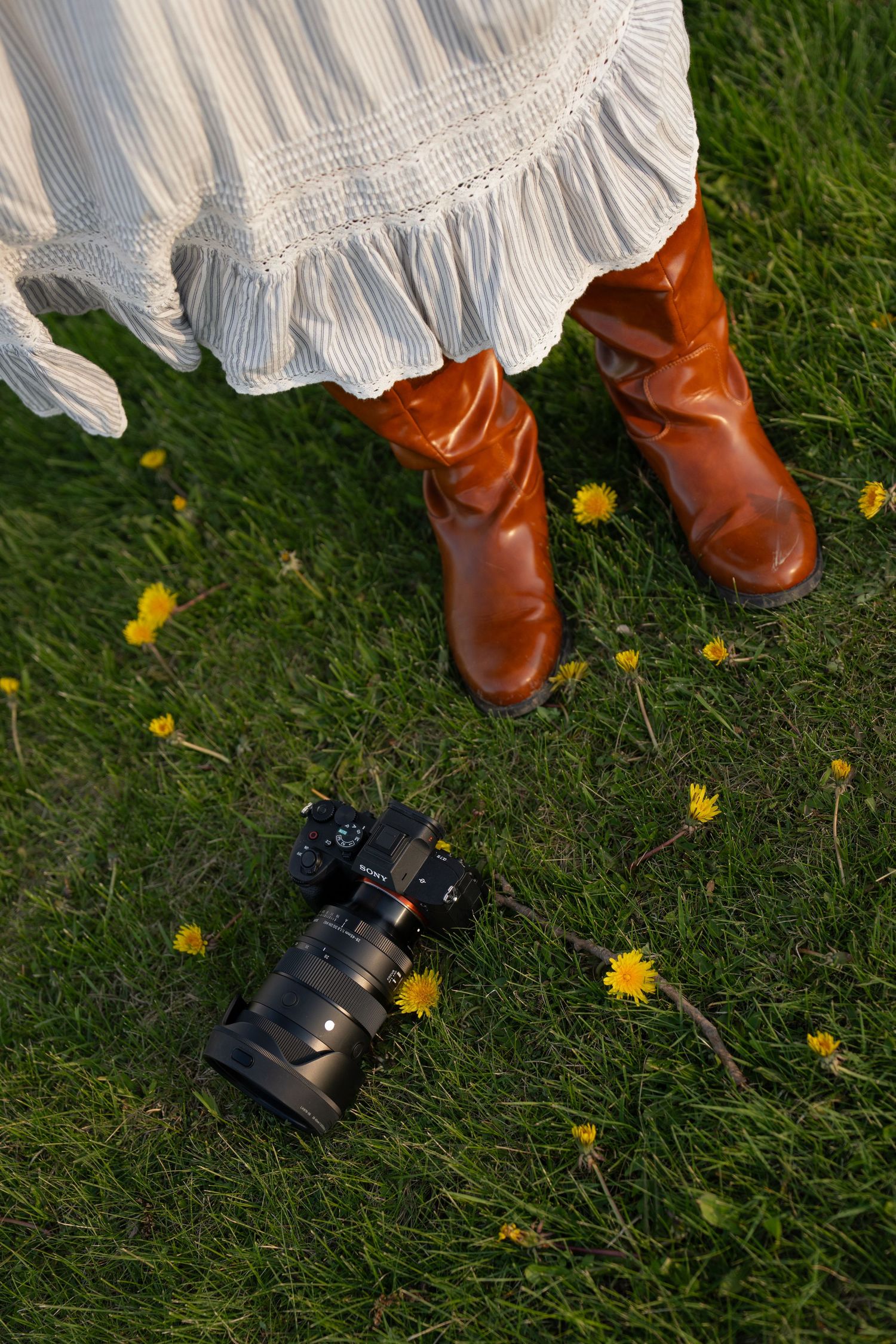 Close-up of woman in white ruffled skirt and brown leather boots standing beside camera on grass.