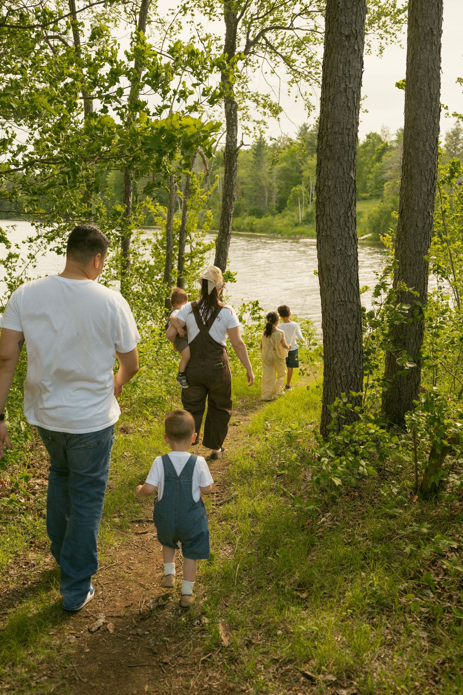 Family with young child walking along wooded riverside trail surrounded by lush green trees in summer.