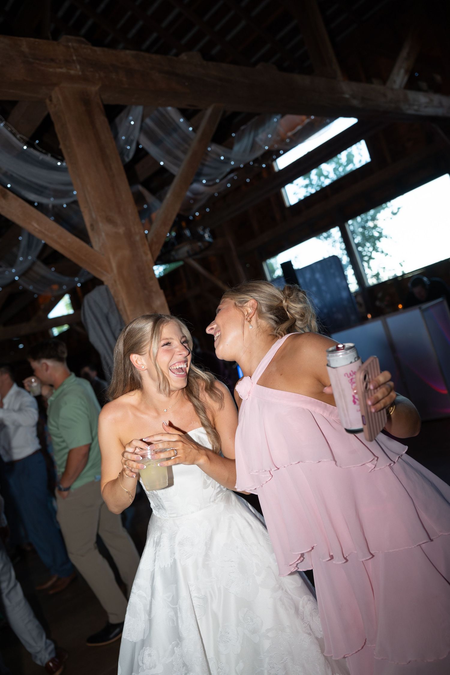 Laughing bride in white dress dancing with bridesmaid in pink dress holding canned drinks at rustic barn reception.