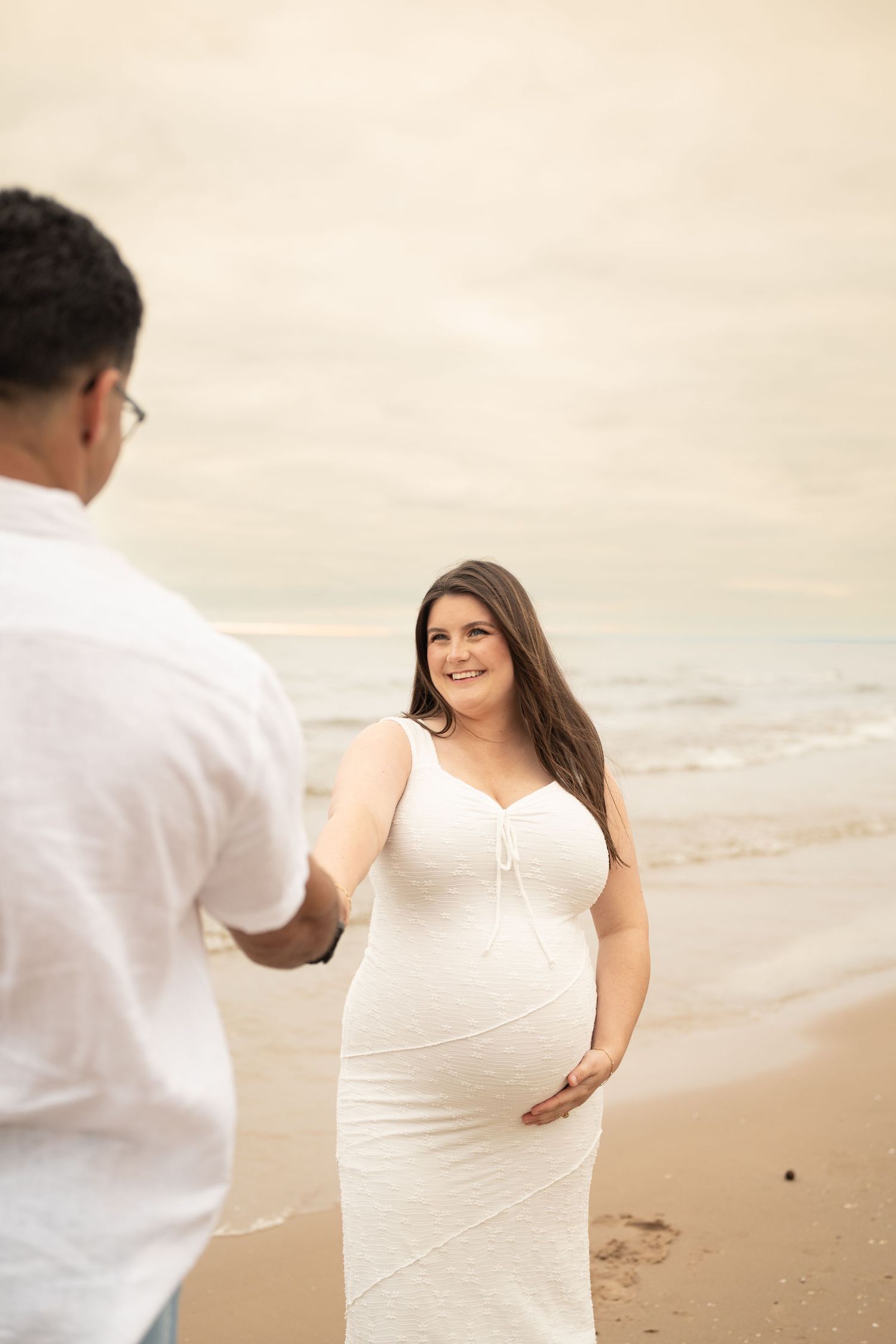 Pregnant woman in white dress holds her belly on a beach while holding hands with her partner.