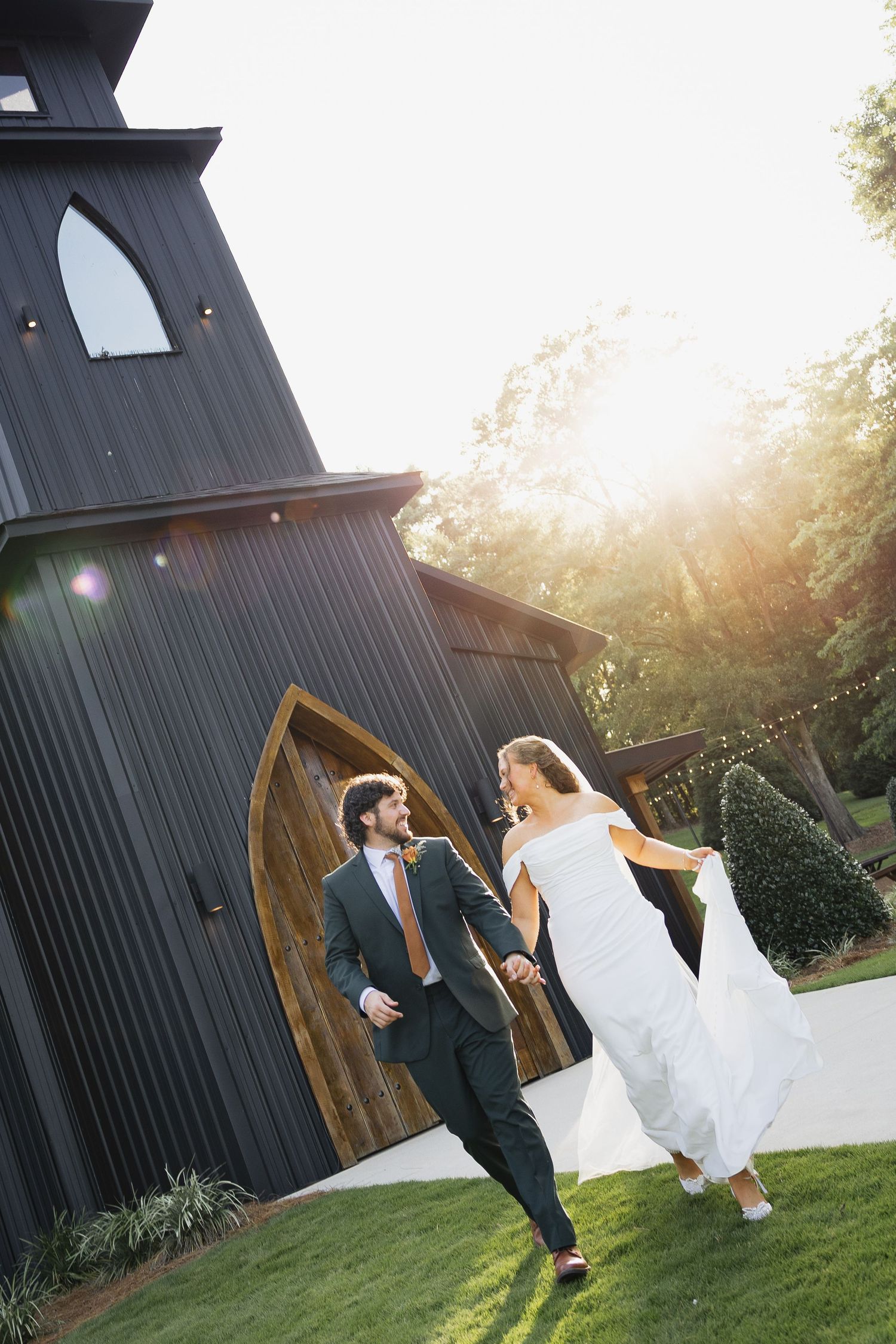 Bride and groom walking joyfully outside a dark barn chapel in golden sunlight.