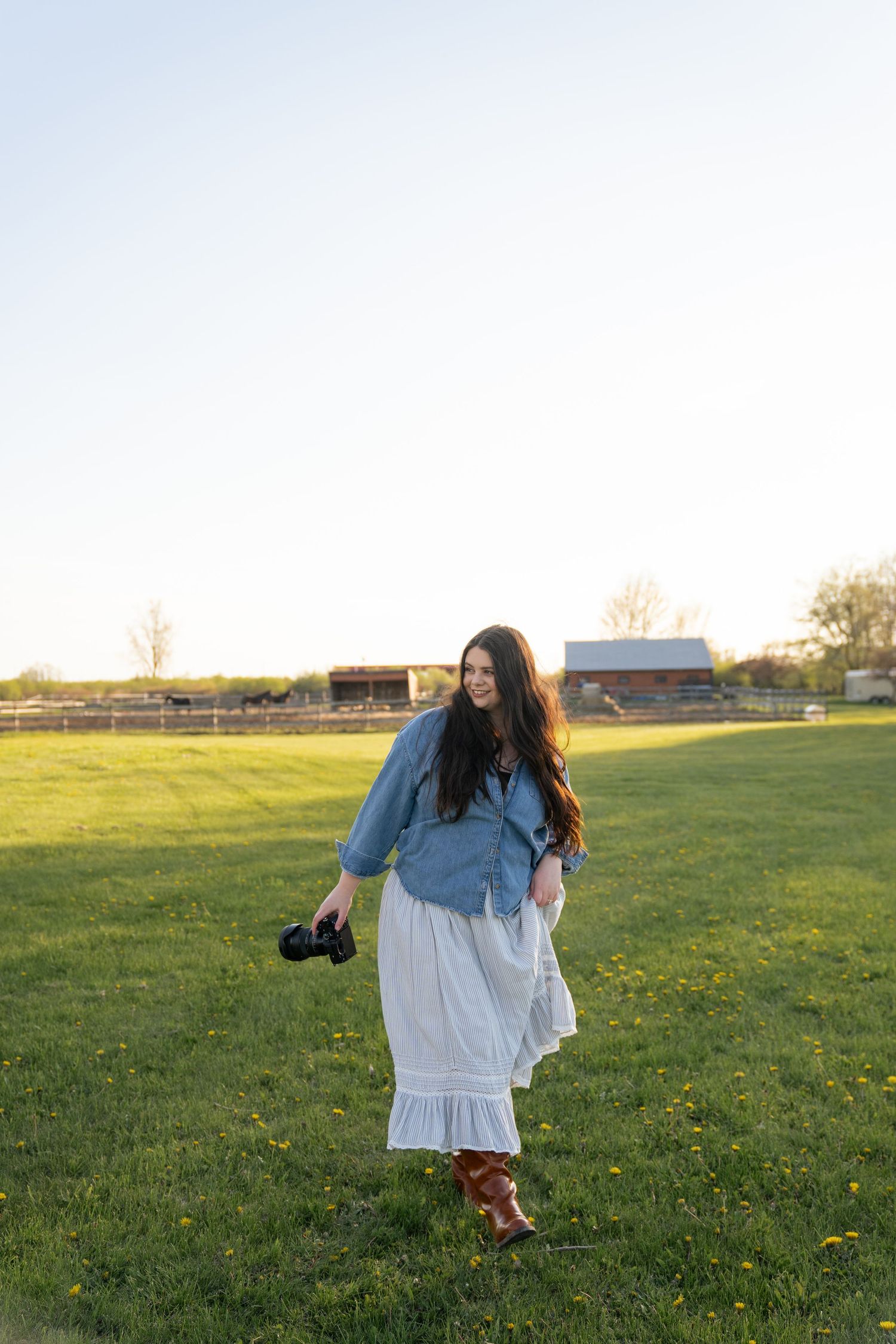 Woman in denim shirt and white maxi skirt holding camera walking through green field at golden hour near rustic barn.