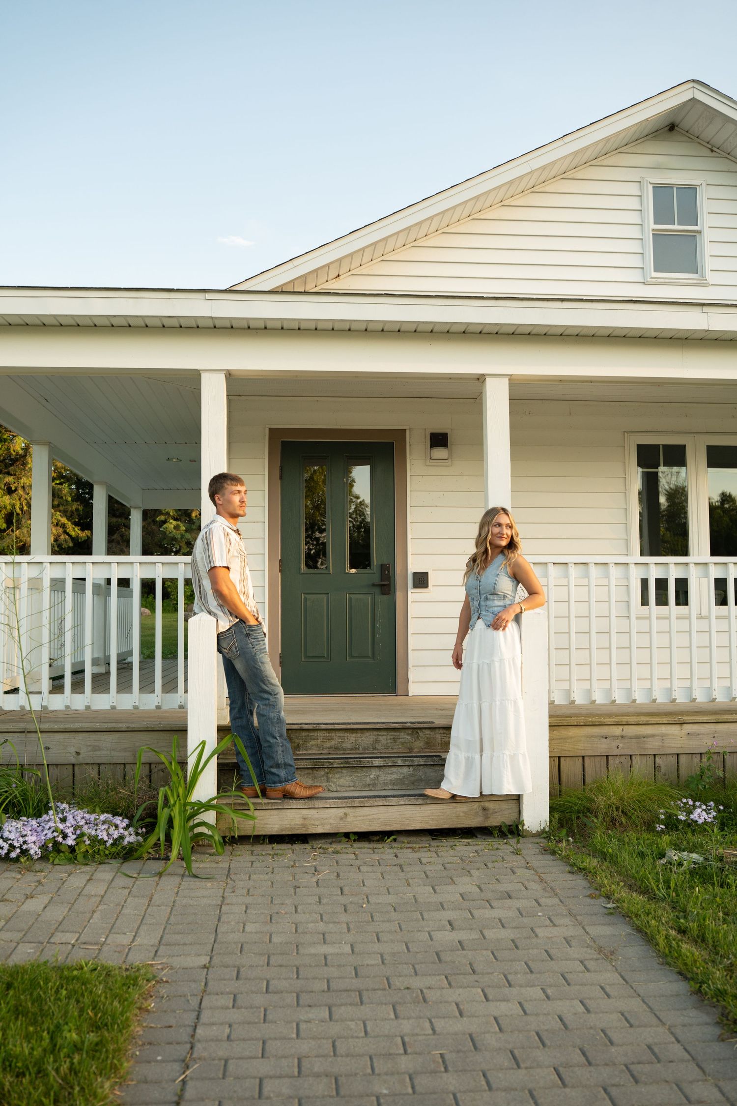Couple standing on porch of white farmhouse at golden hour, man in jeans and woman in white skirt.