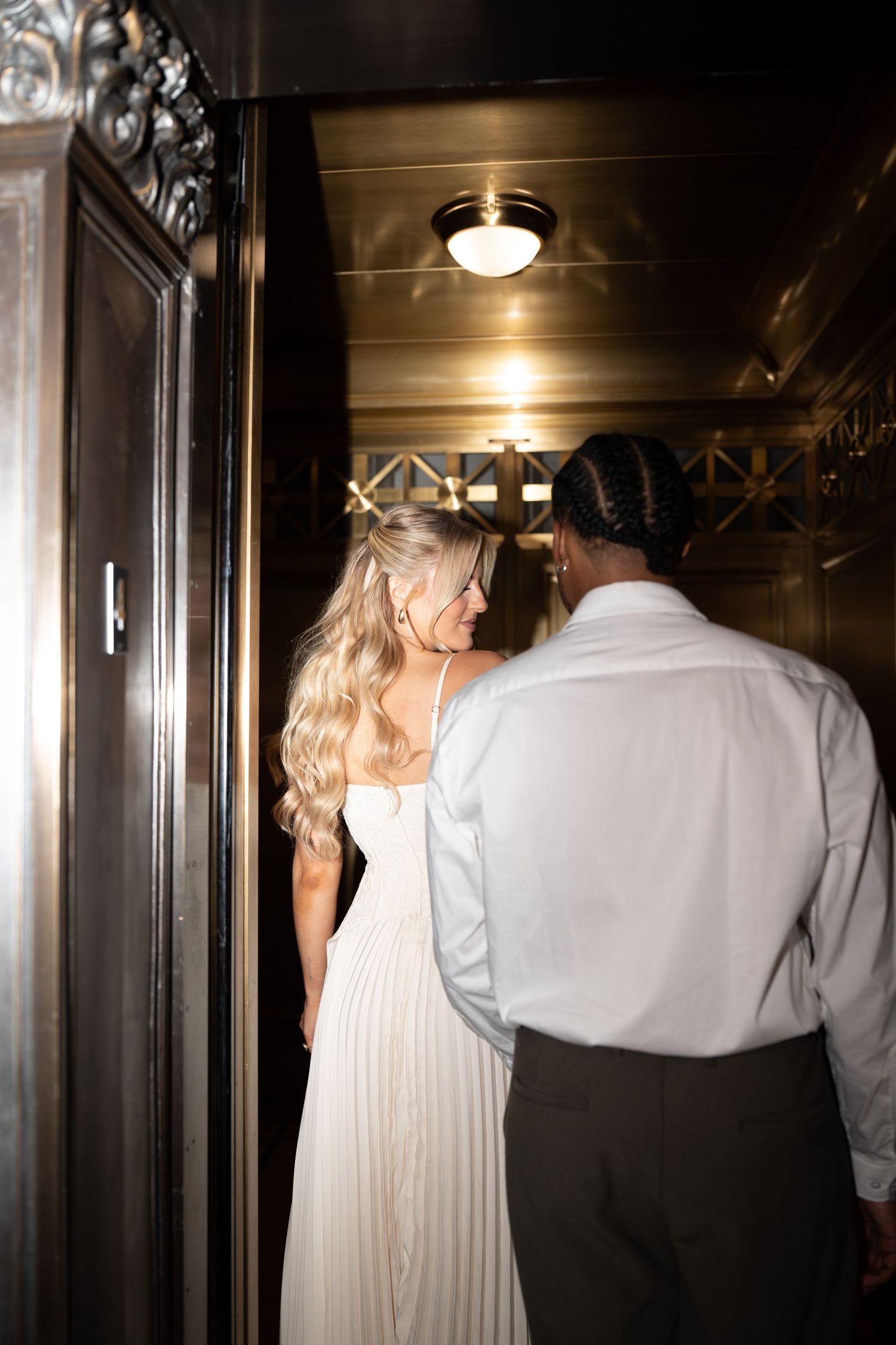 Bride in white gown and groom in white shirt entering elegant elevator with ornate gold interior lighting.
