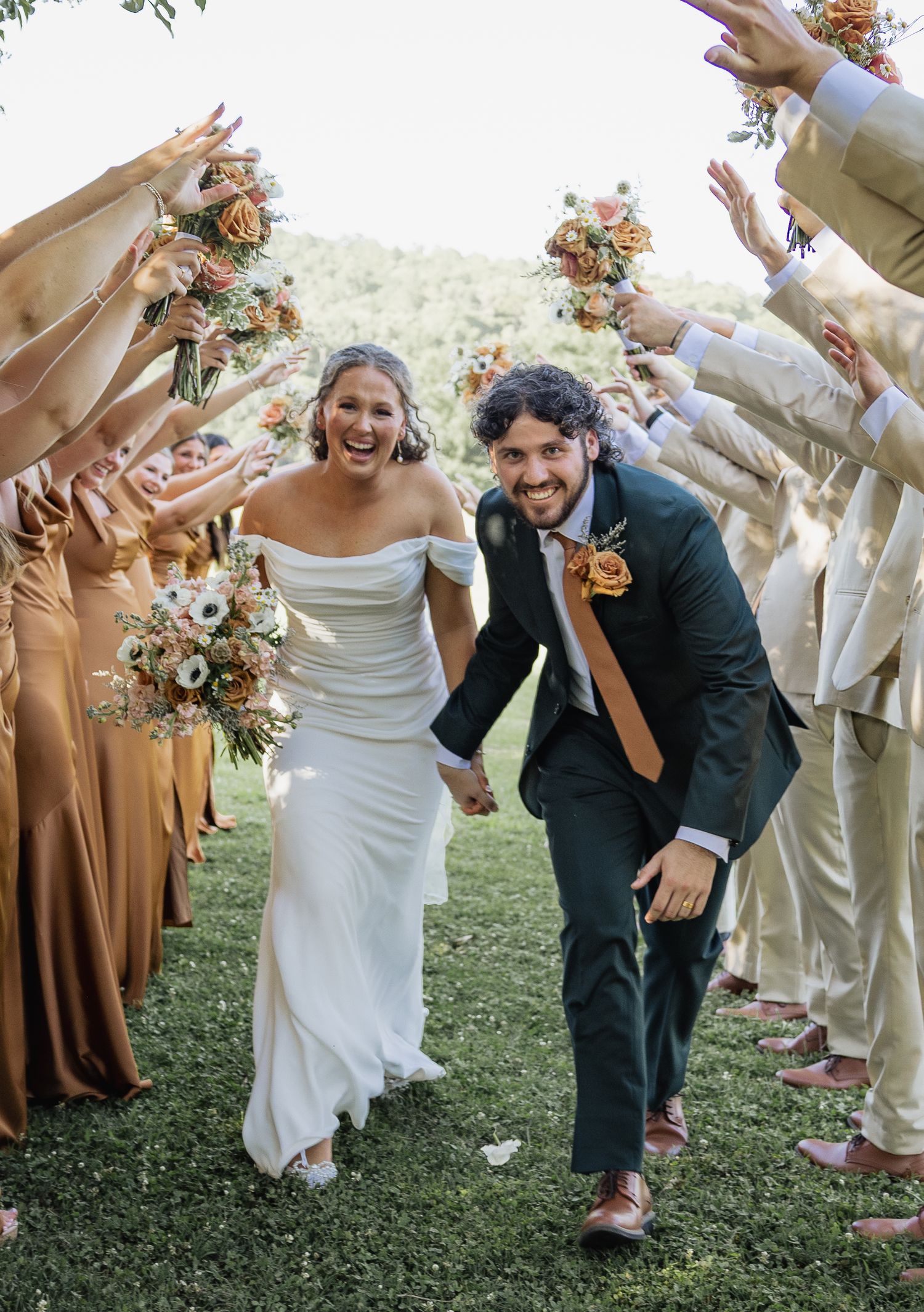 Joyful newlyweds running through arch of raised hands formed by wedding party in golden autumn setting.