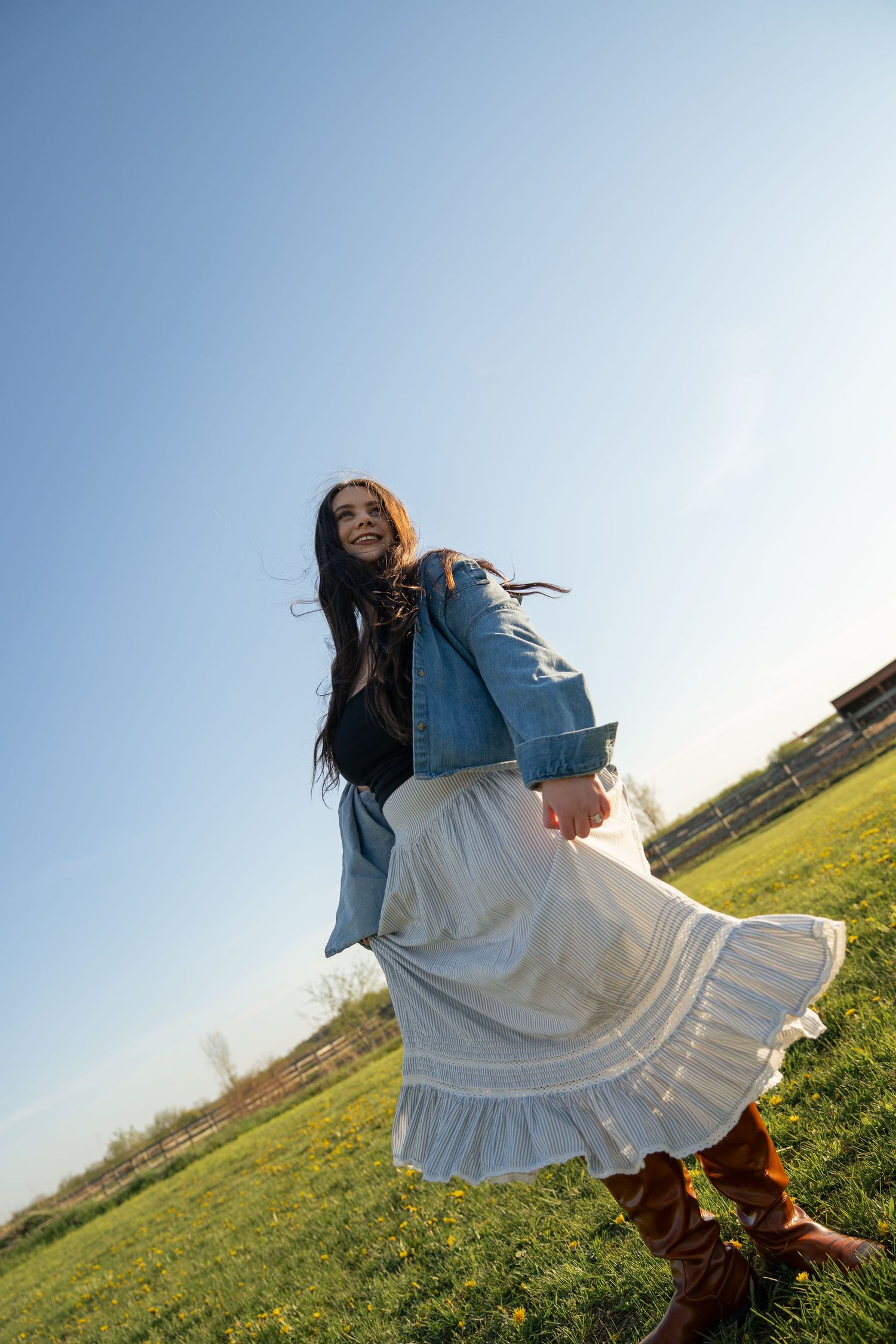 Young woman in denim jacket and white boho skirt twirling in a sunny green field under blue sky.