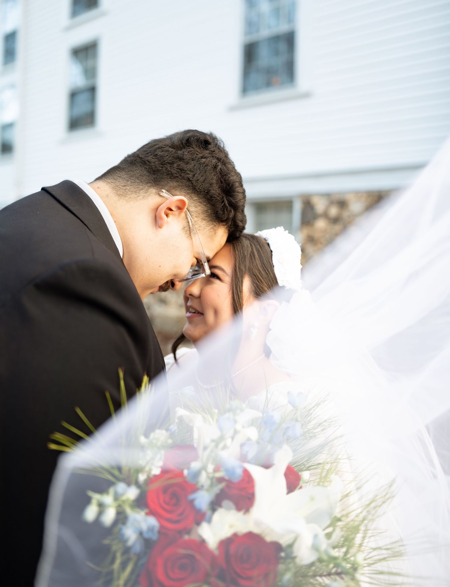 Groom in black suit leaning toward smiling bride holding red rose bouquet under flowing veil outdoors.