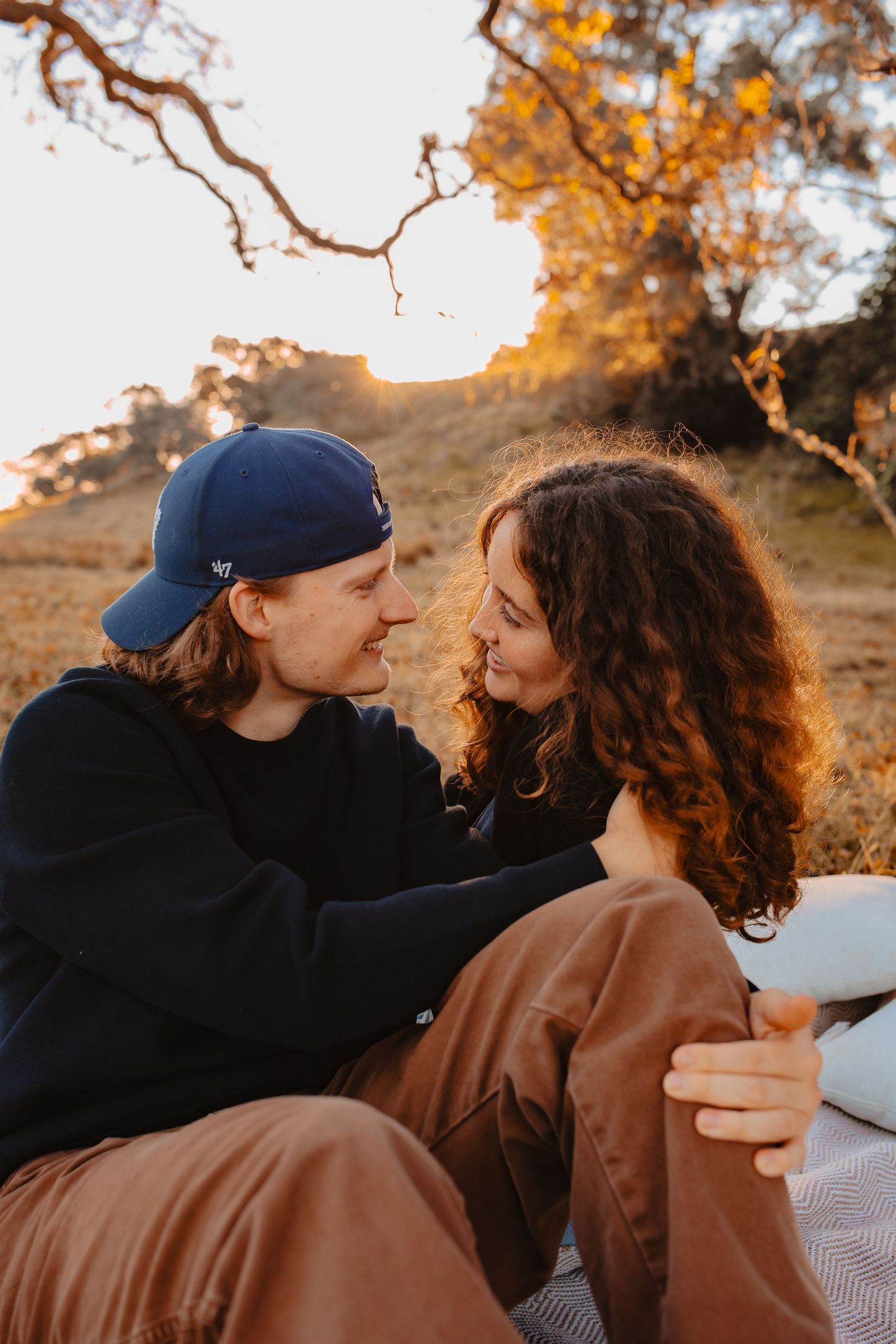 Wide shot of a man and woman sharing an intimate moment under a large, rustic tree during a golden hour outdoor portrait session.