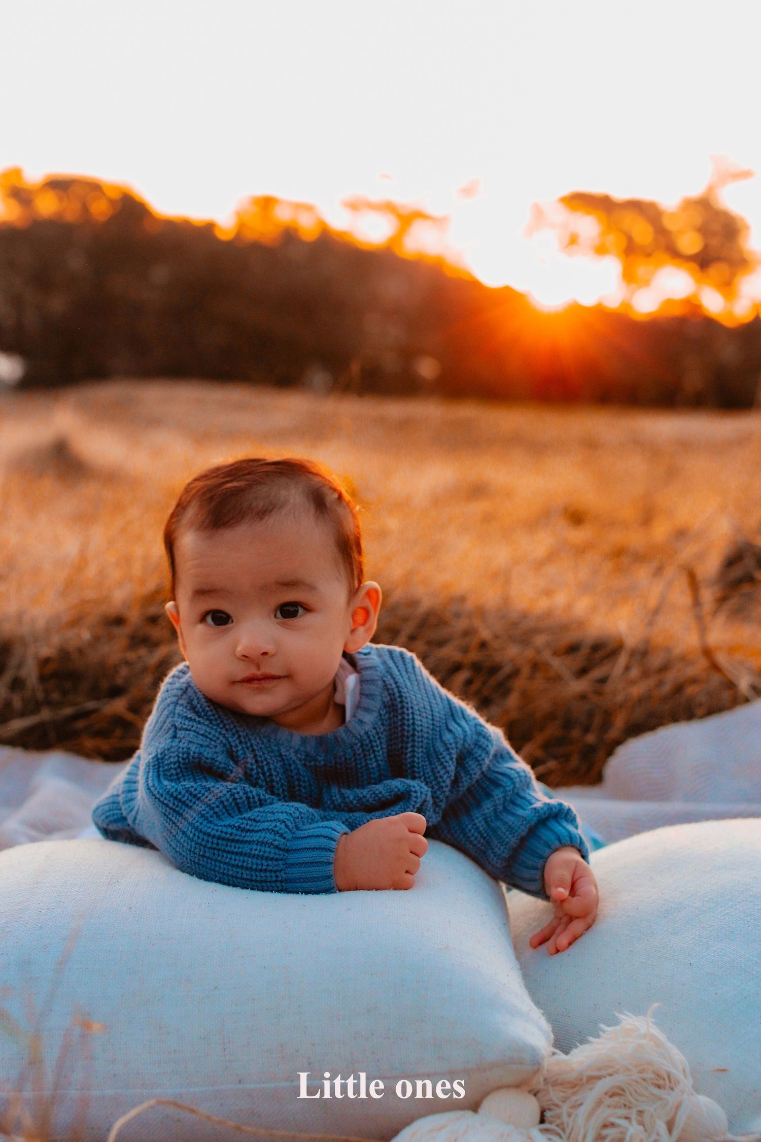 A baby boy wearing a blue knit sweater lying on a white cushion outdoors during a vibrant golden hour sunset at One Tree Hill, Cornwall Park