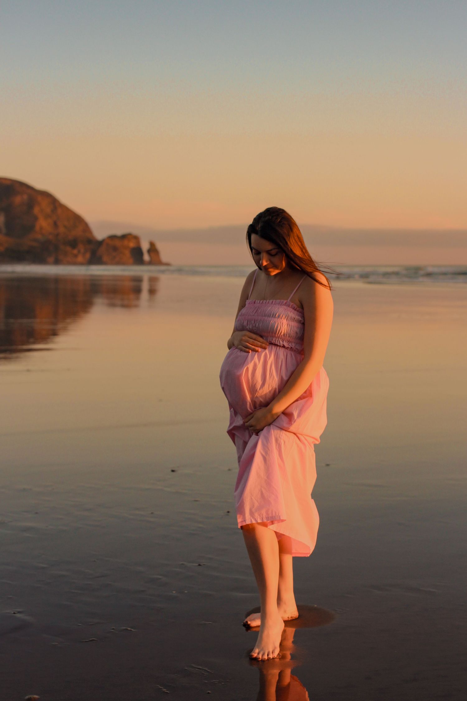 Close-up of a pregnant woman’s belly held by hands in a white lace dress, backlit by warm golden hour light.