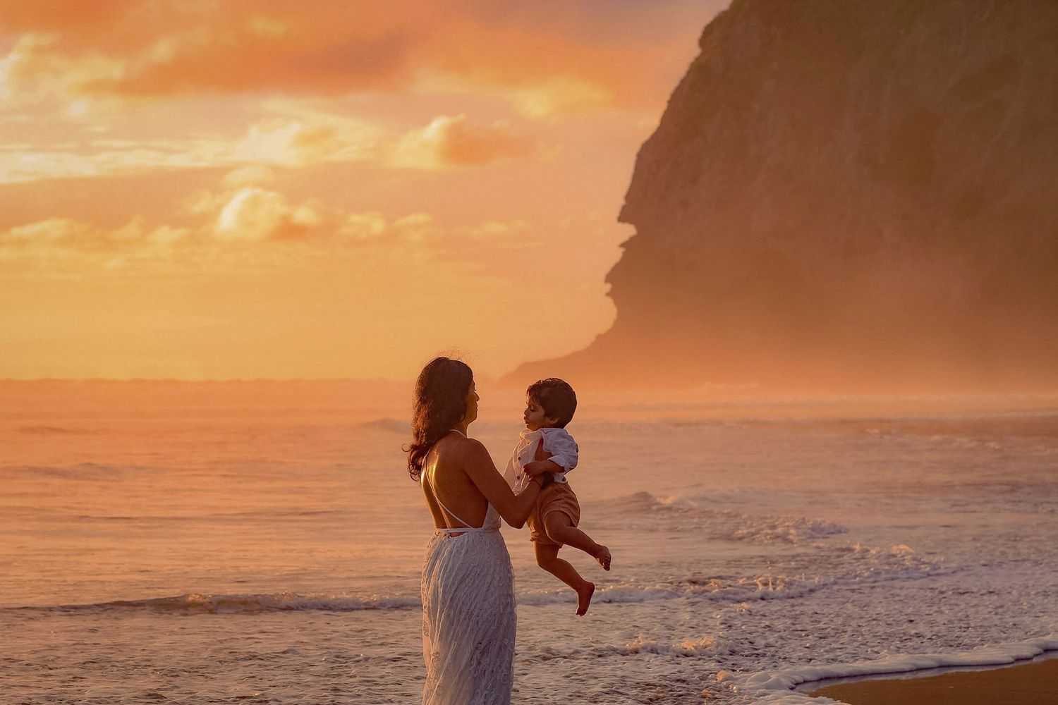 Mother playing with her baby with a bright sunset in the background at piha, with the water mist and Lions Rock in the background.