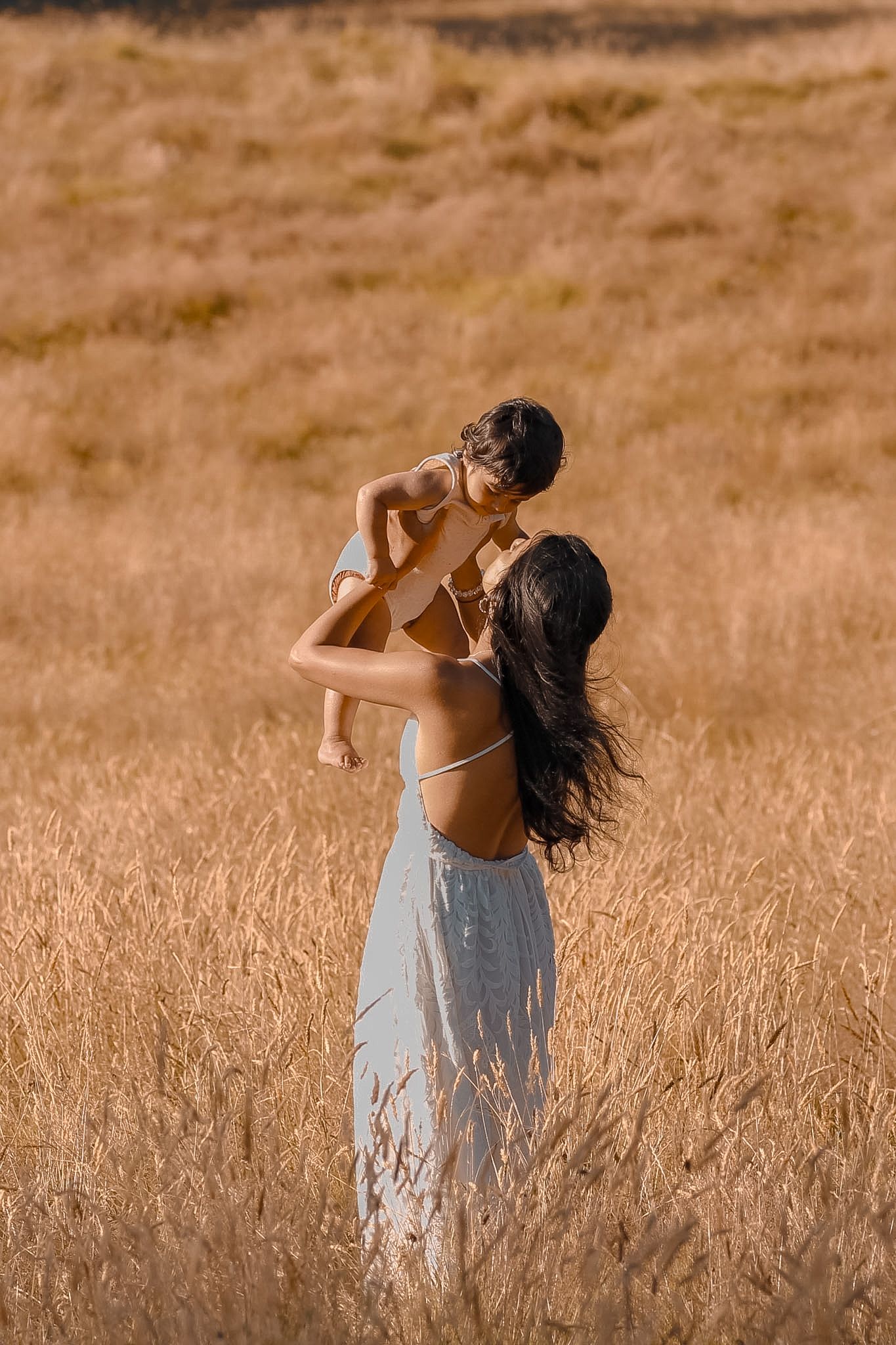 Soft-focus portrait of a mother and child in a meadow with her hair moving in the soft wind and baby laughing and looking back at his mum.