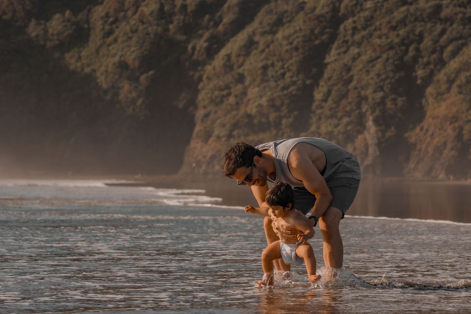 Baby walking into the low tide in nappy, photo captures interaction between father and son.  Photo of emotional candid moments