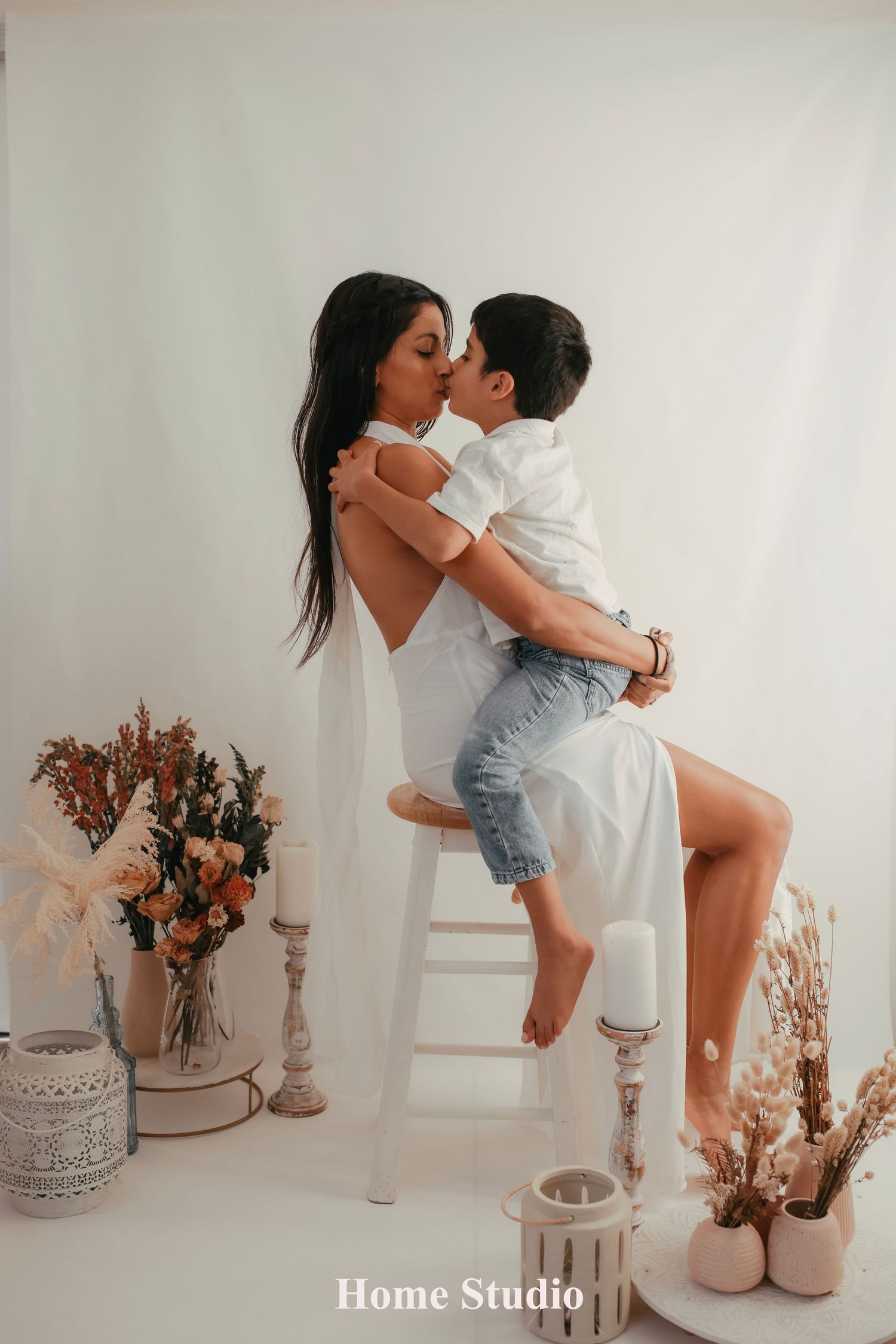 A mother in a white dress kissing her young son while sitting on a stool during an Auckland home studio photography session.