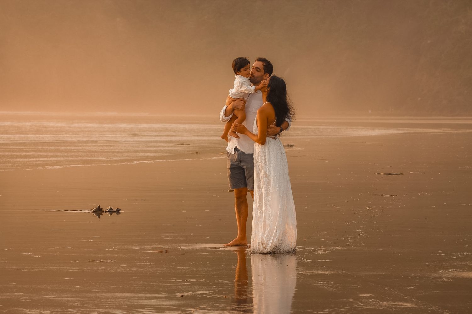 Dad giving a kiss to his toddler while holding his wife. Golden hour photographer based in auckland.