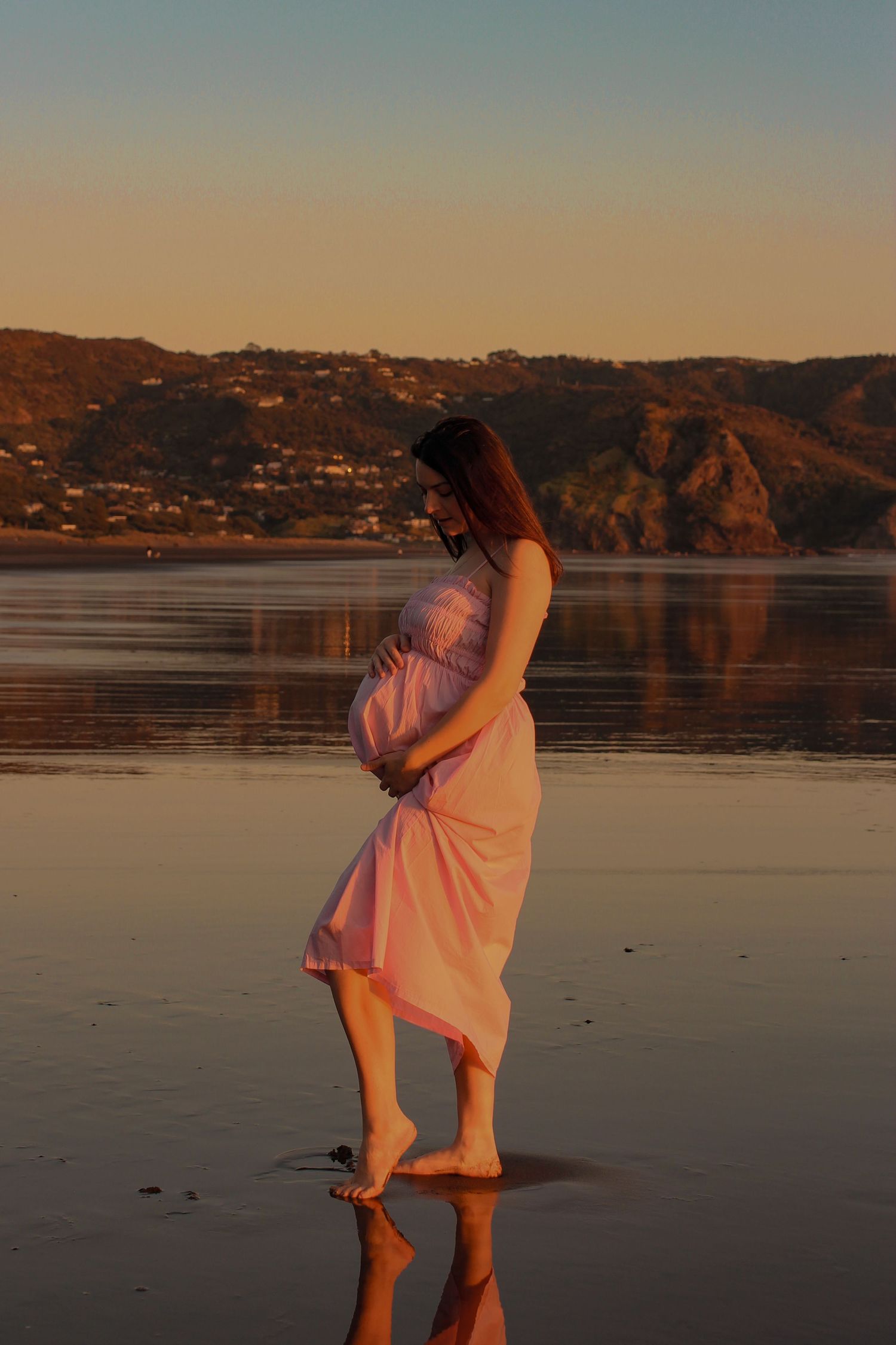 Close-up of a pregnant woman’s belly held by hands in a pink lace dress, backlit by warm golden hour light.