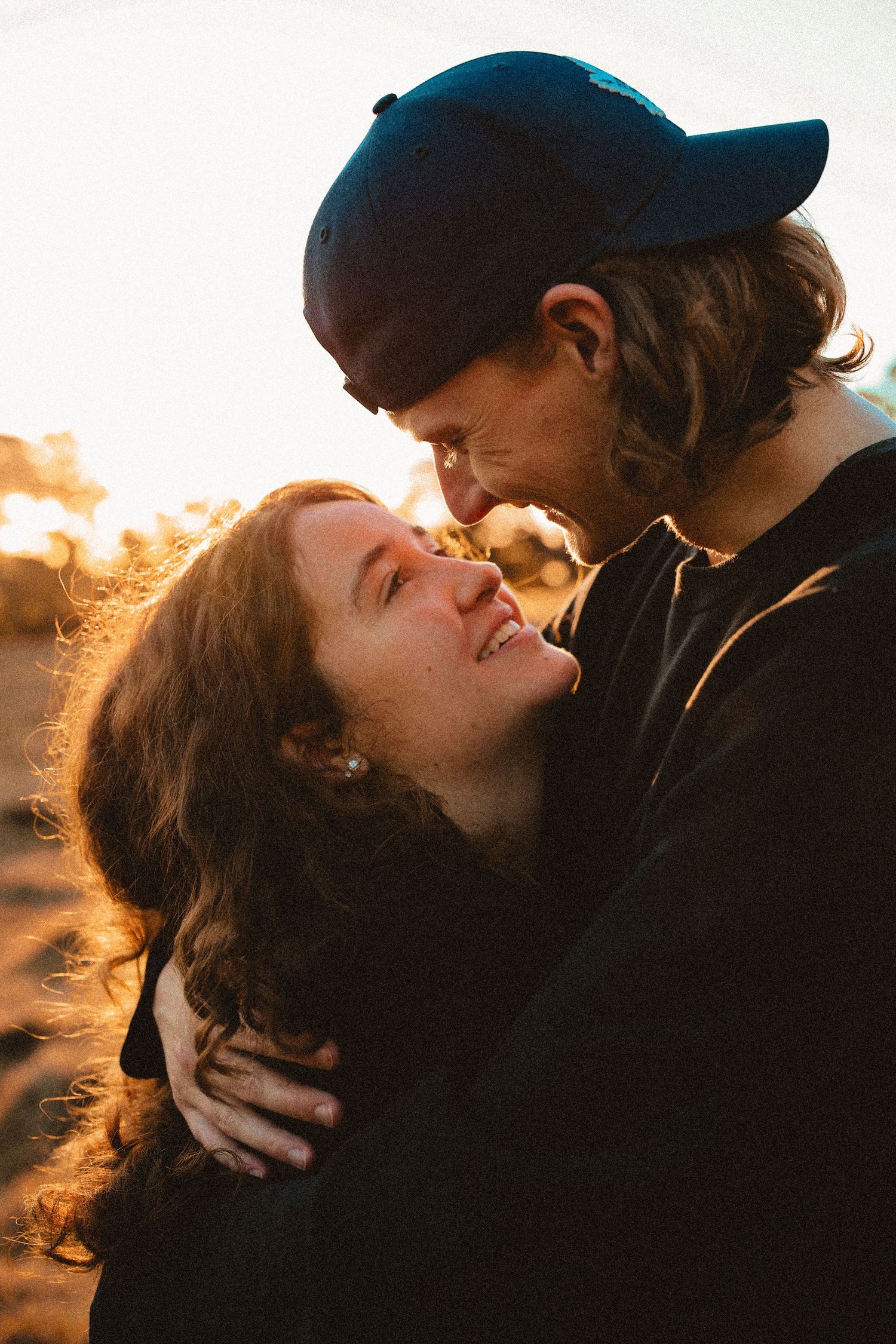 Close-up portrait of a man tenderly hugging a woman from behind in an outdoor field at sunset.