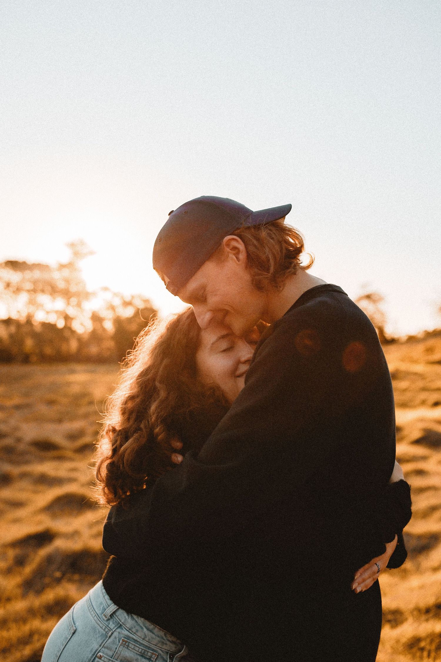 A couple smiling and walking hand-in-hand through a sunlit field during a golden hour engagement photoshoot.