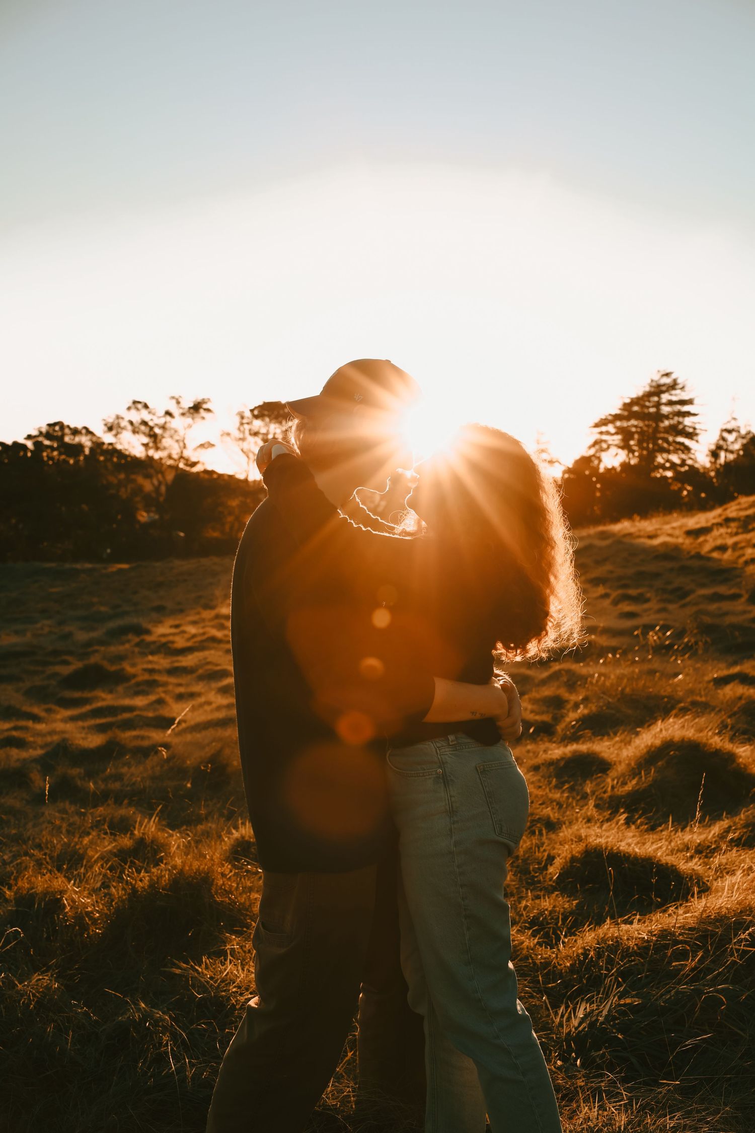 A woman with long curly hair hugging a man wearing a backward cap in a field.
