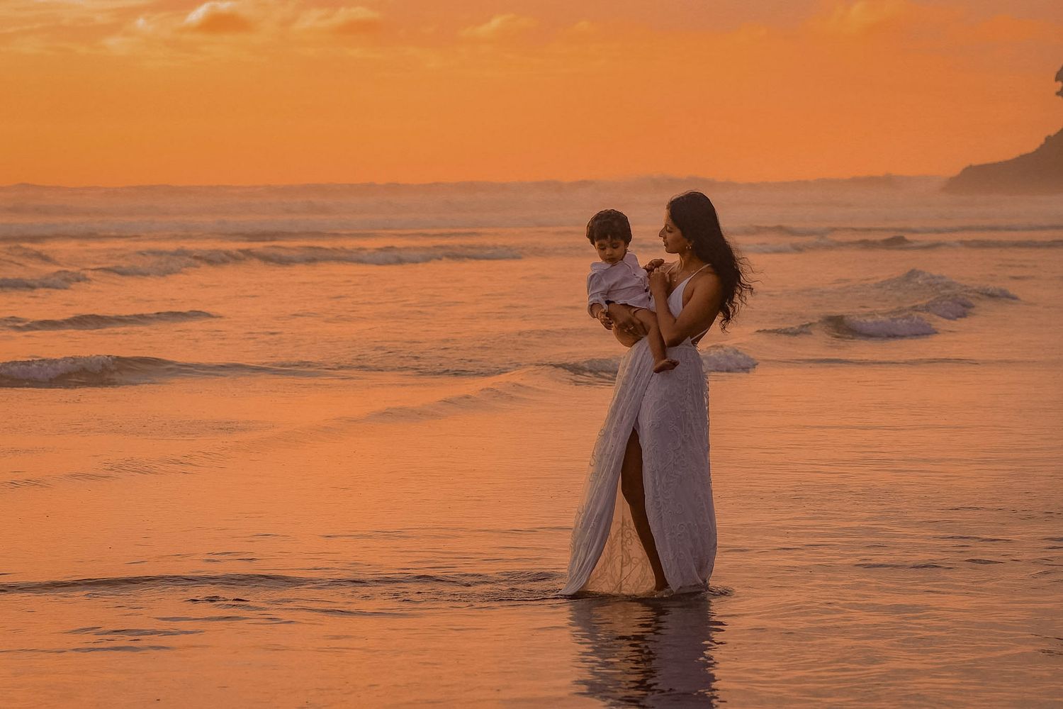 Mum walking into the water with her baby, in a boho lace dress with the sun in the background setting. Sky painted golden and orange.
