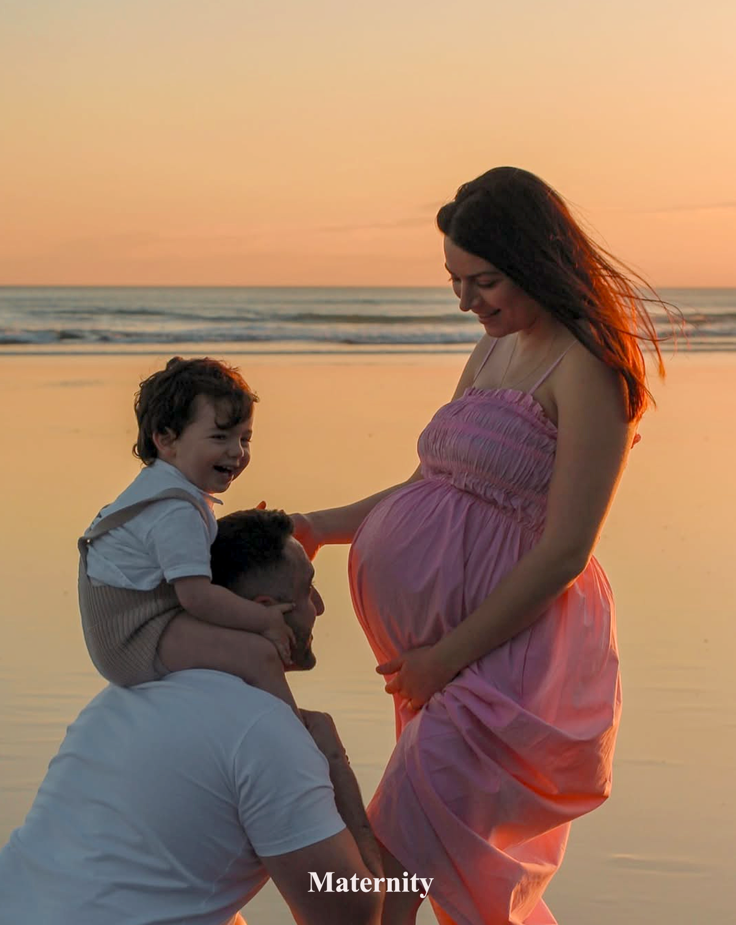 A dad carrying his child on his shoulders, while kissing this wifes tummy, a sunset photography session at Piha. Maternity photography