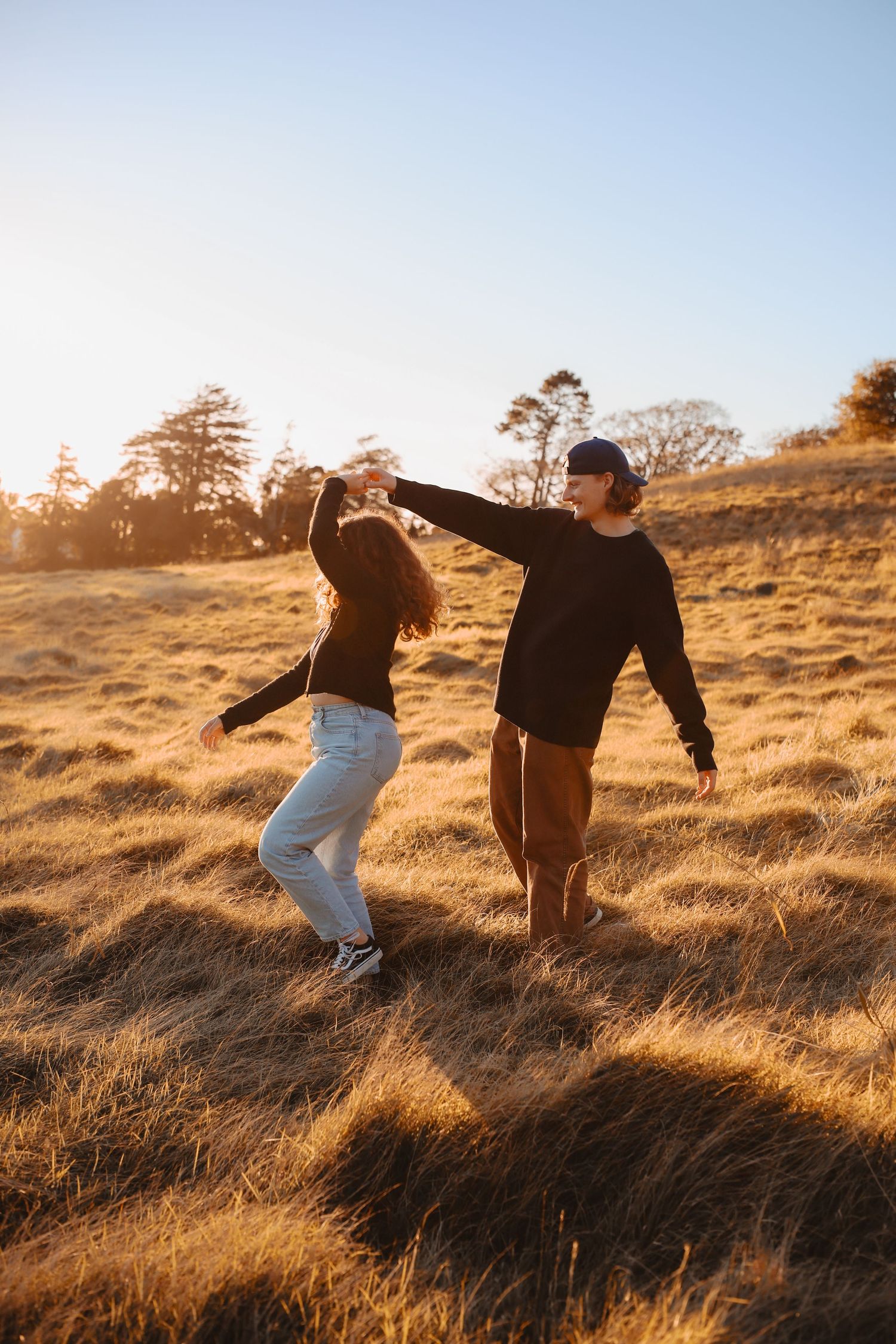 A young man and woman dancing together in a tall grass field with the bright evening sun setting behind them.