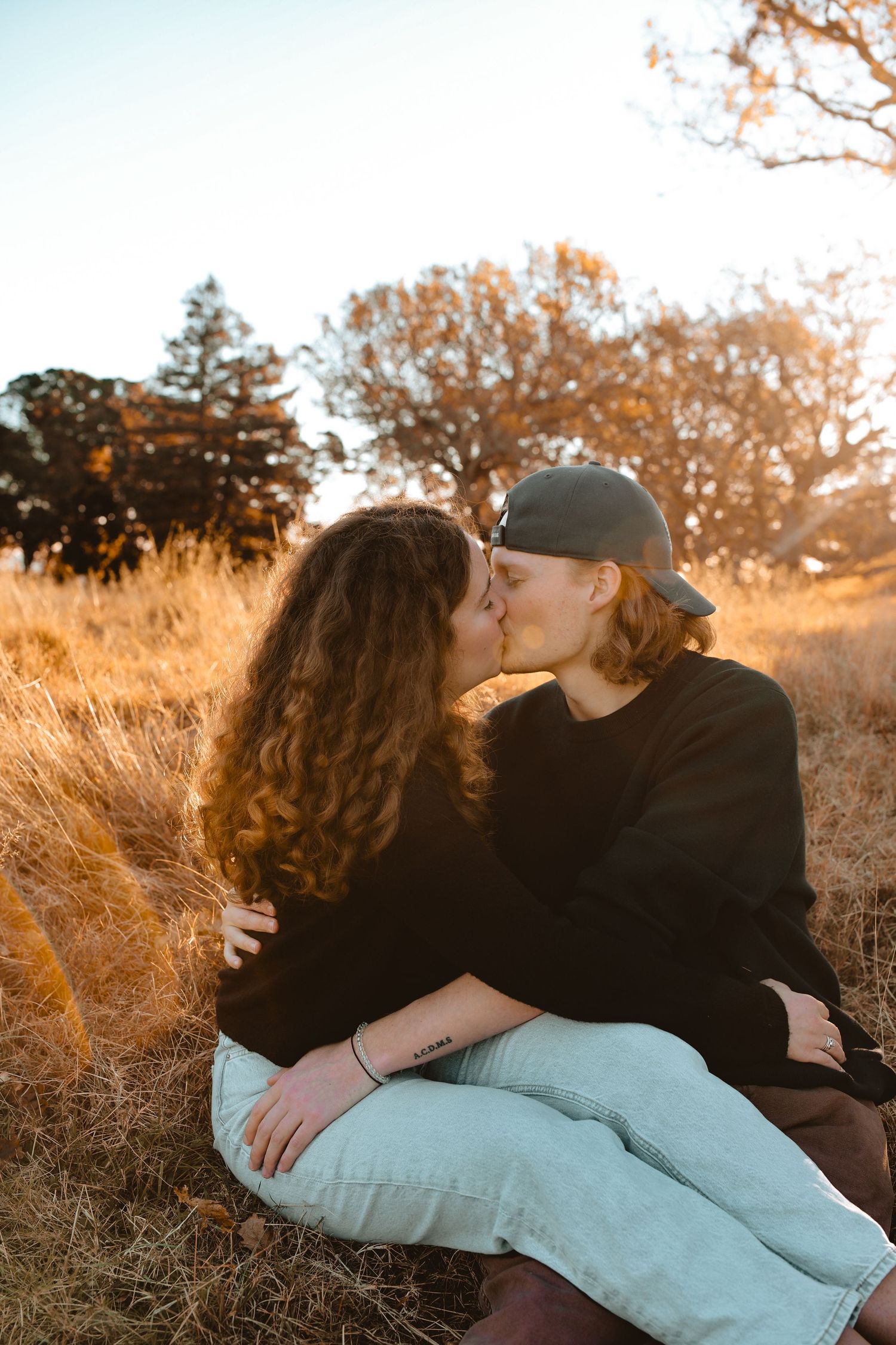 A man and woman sitting side-by-side on a white blanket in a rustic meadow, backlit by the warm, soft light of a setting sun.