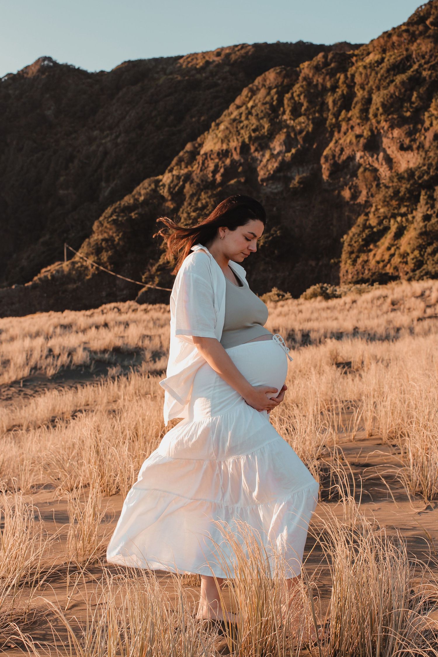 Sunset maternity photography session at Piha Beach, with the soft wind flowing her long hair and lace white dress.