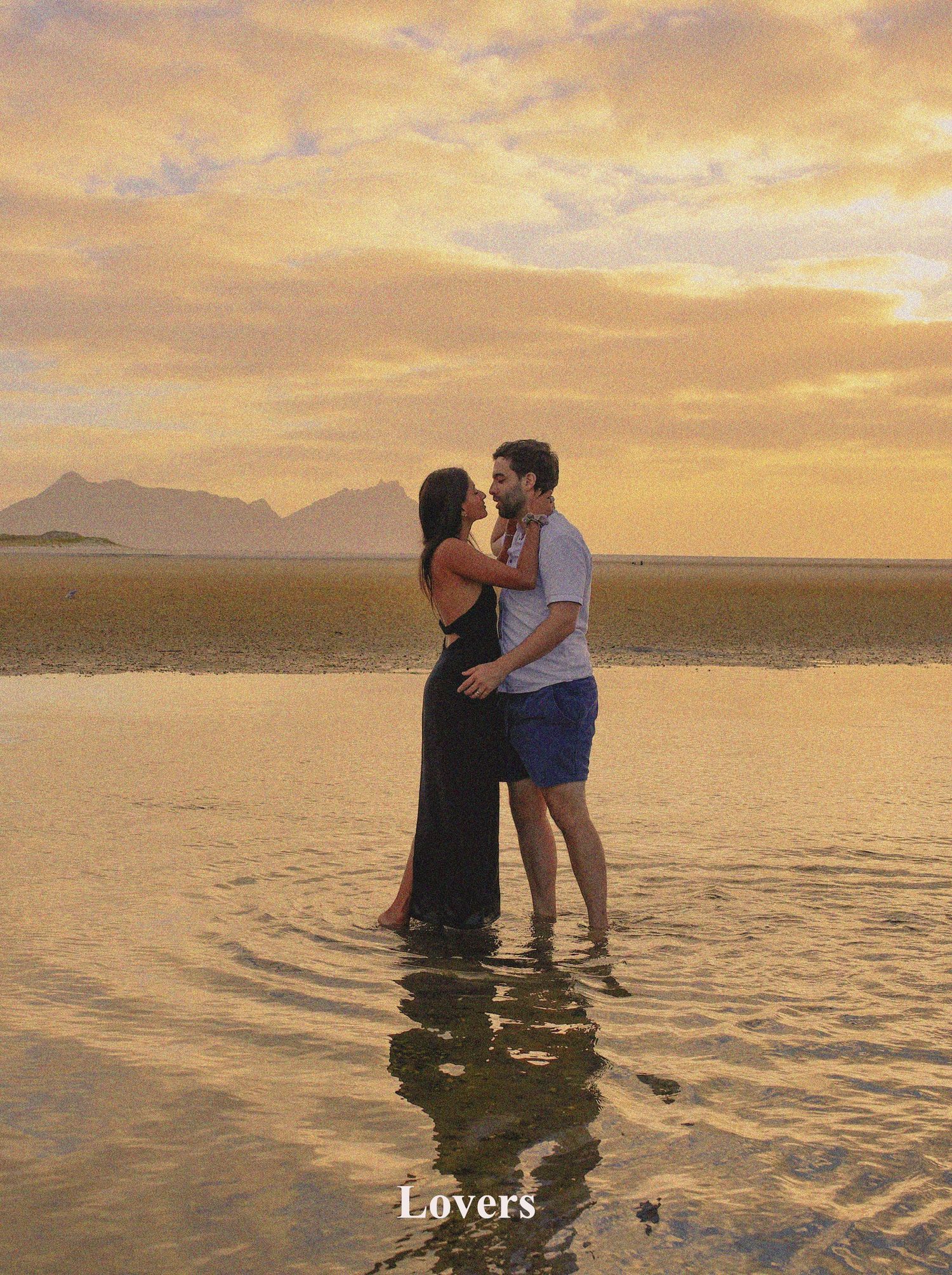 A romantic couple embracing while standing in the shallow water during a golden hour sunset beach session in Auckland. Ruakaka Beach