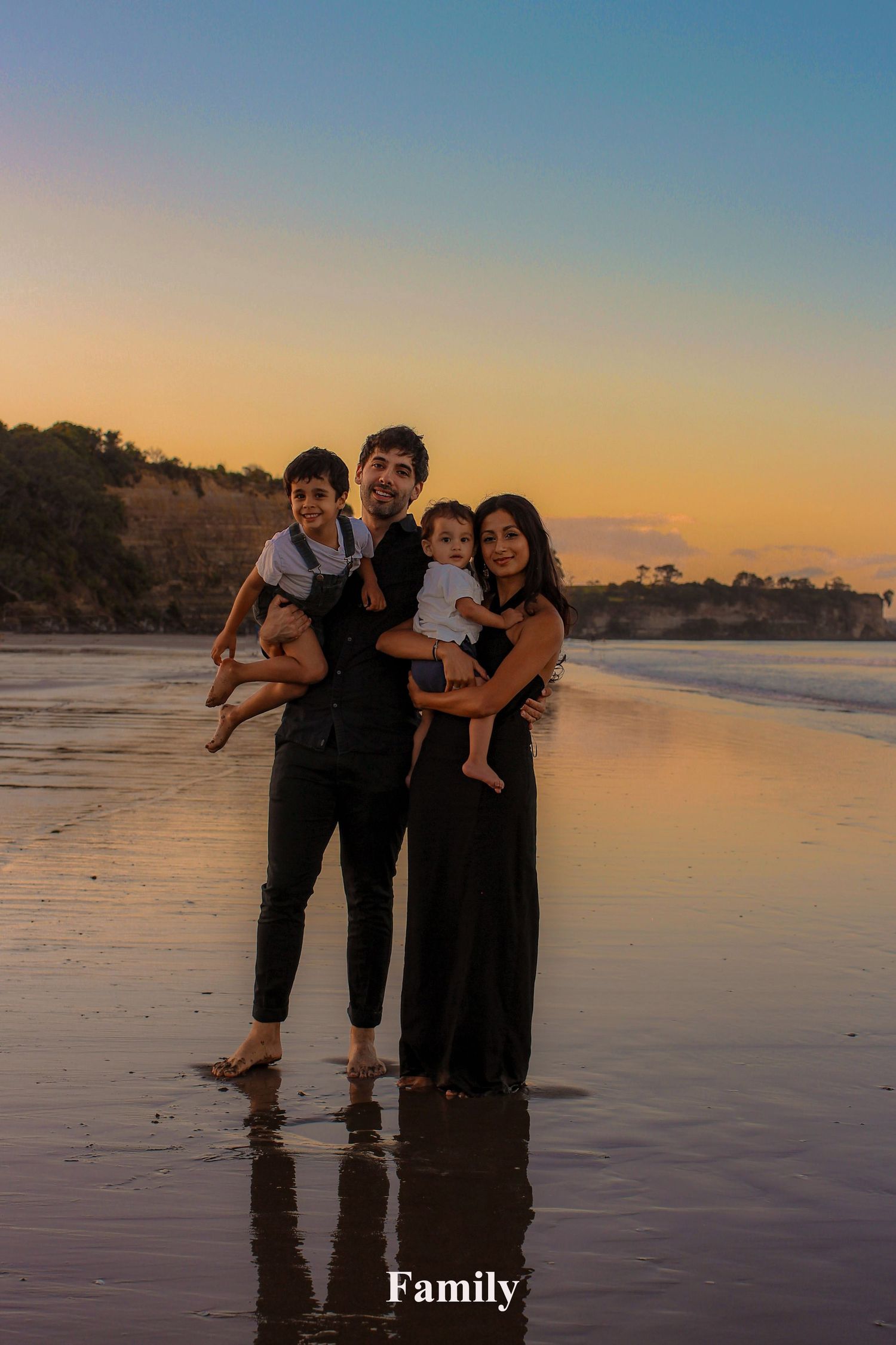 A family of four standing together in the shallow water at an Auckland Long Bay beach during a golden hour sunset portrait session.