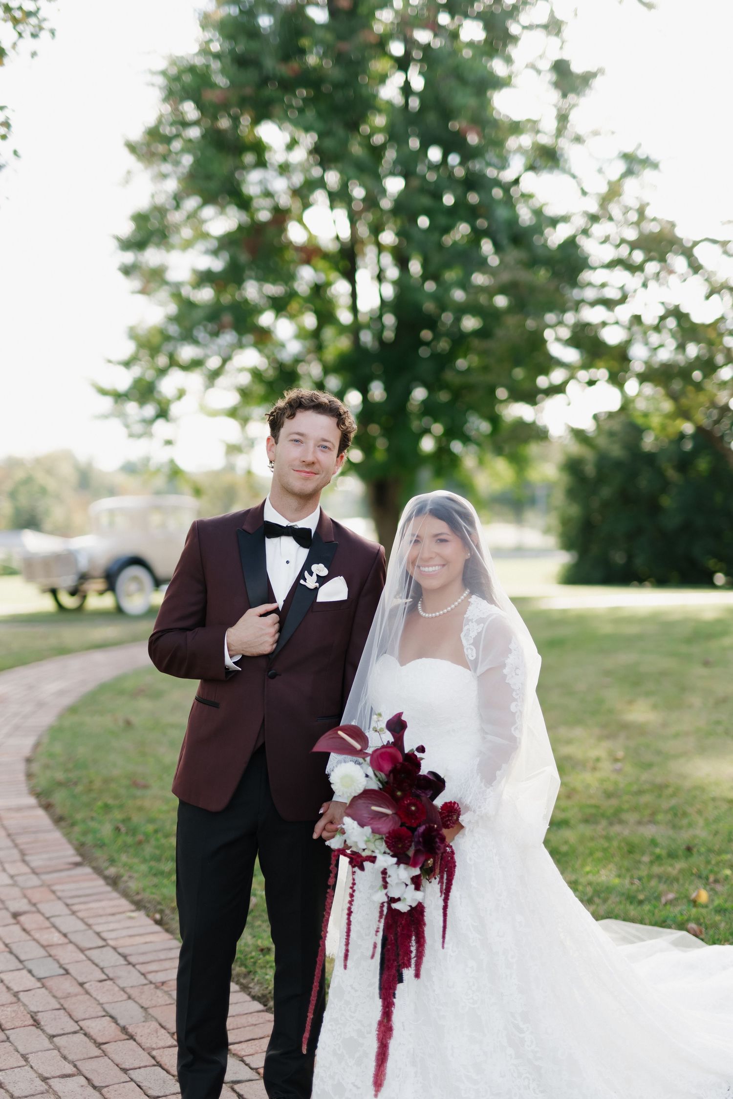Bride and groom pose on brick path, groom in burgundy tuxedo, bride in lace gown with veil and red floral bouquet.