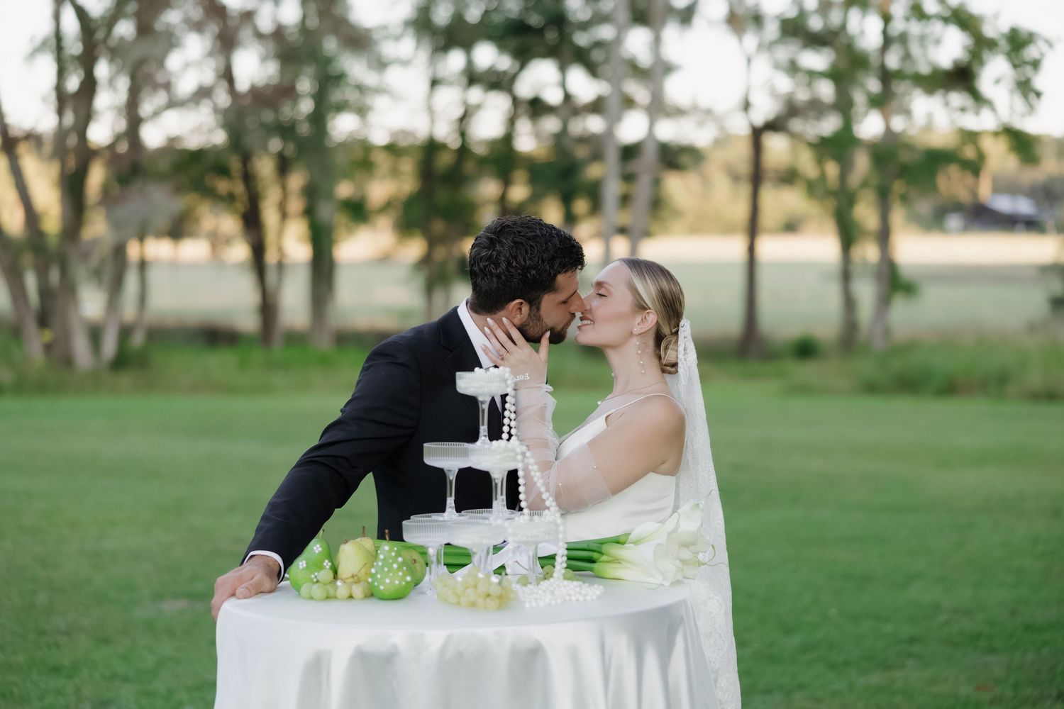 Bride and groom sharing a kiss beside their white wedding cake outdoors on a lush green lawn.
