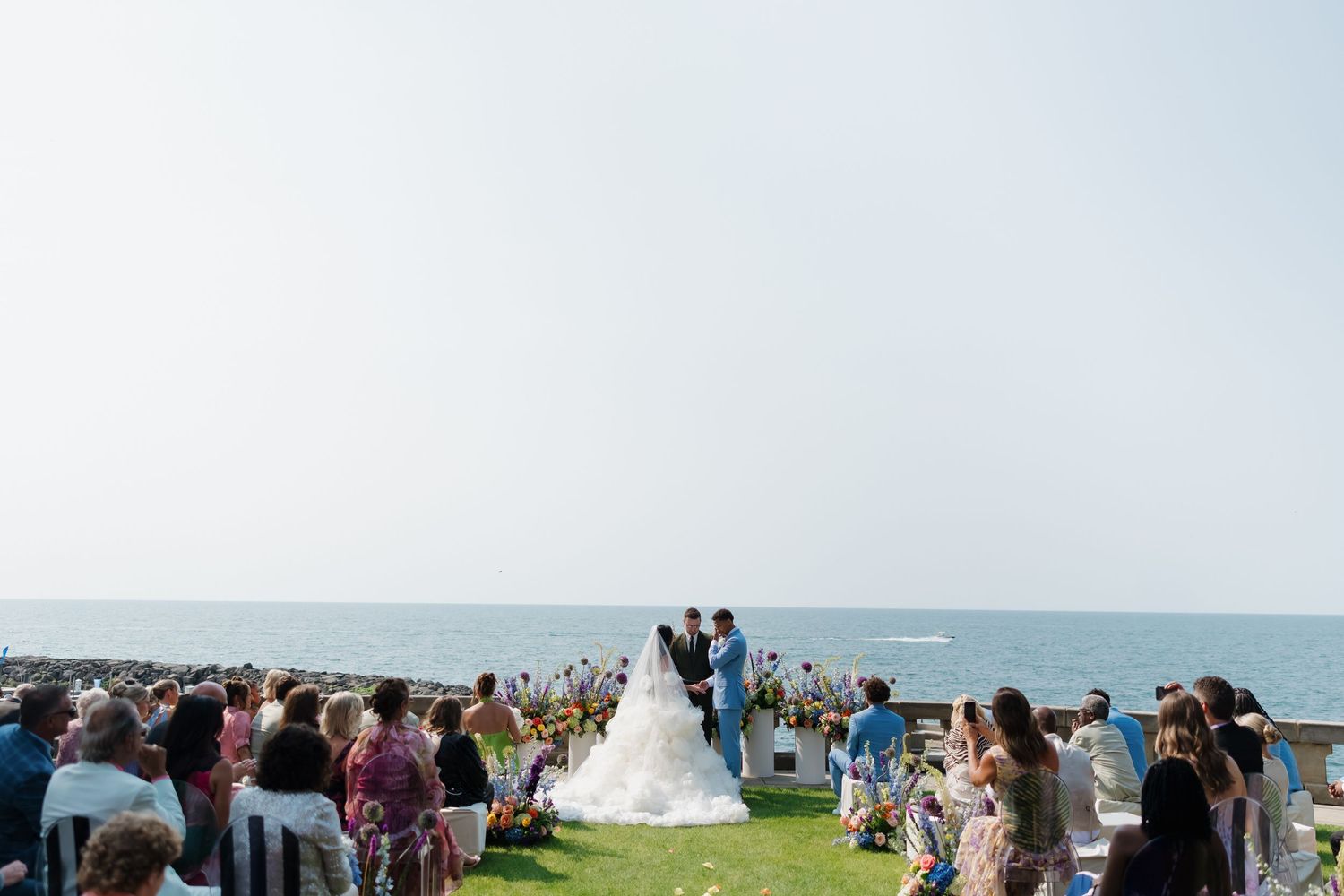 Outdoor wedding ceremony overlooking the ocean with bride and groom at altar surrounded by seated guests on a sunny day.
