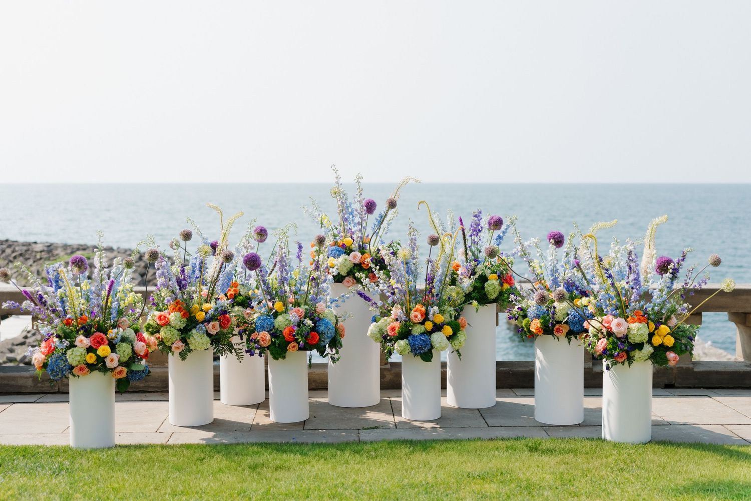 Colorful wildflower arrangements in white vases displayed outdoors by the ocean at a seaside wedding ceremony.