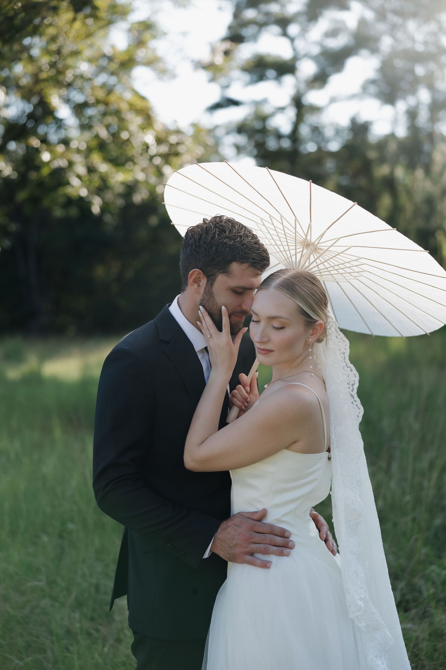 Bride in a white dress and lace veil holds a white parasol while embracing her groom in a dark suit in a sunlit green meadow.