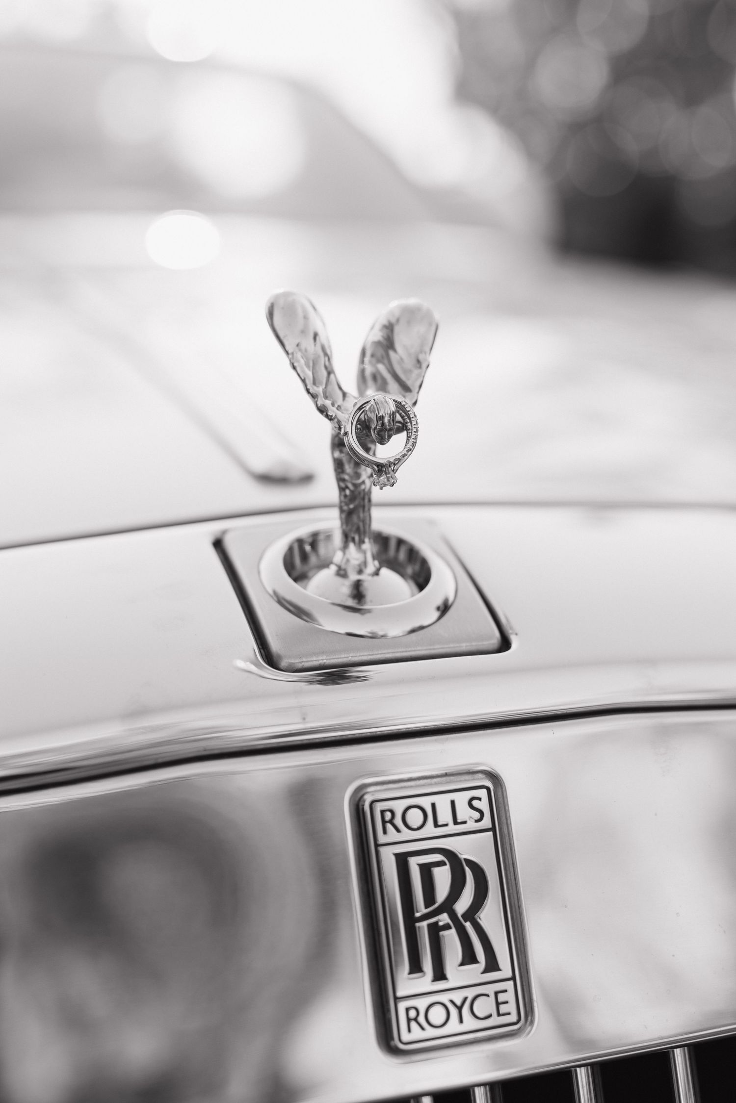 Black and white close-up of Rolls-Royce Spirit of Ecstasy hood ornament and iconic RR badge on luxury car.