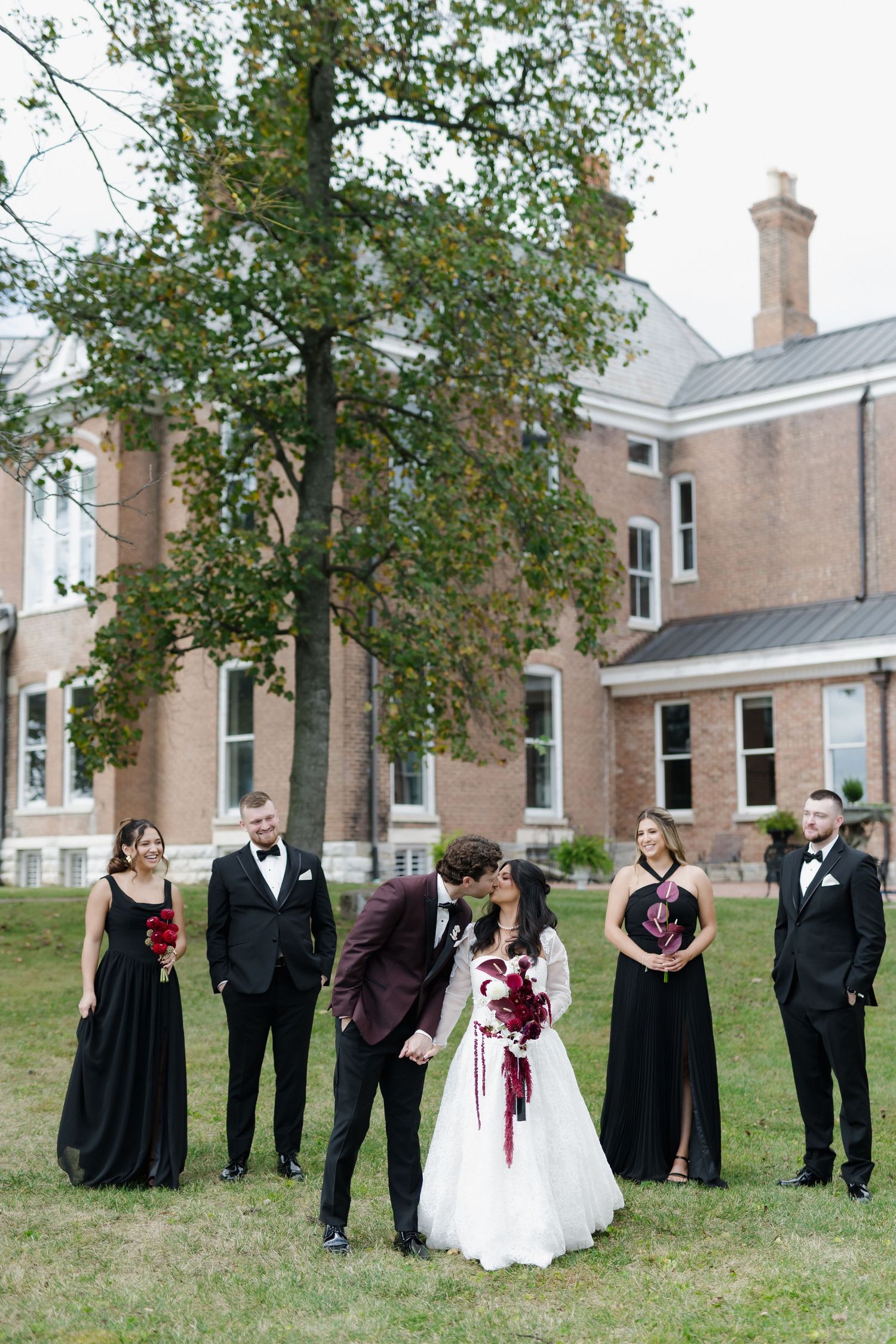 Bride and groom kiss surrounded by wedding party in black attire with red bouquets outside a brick estate.