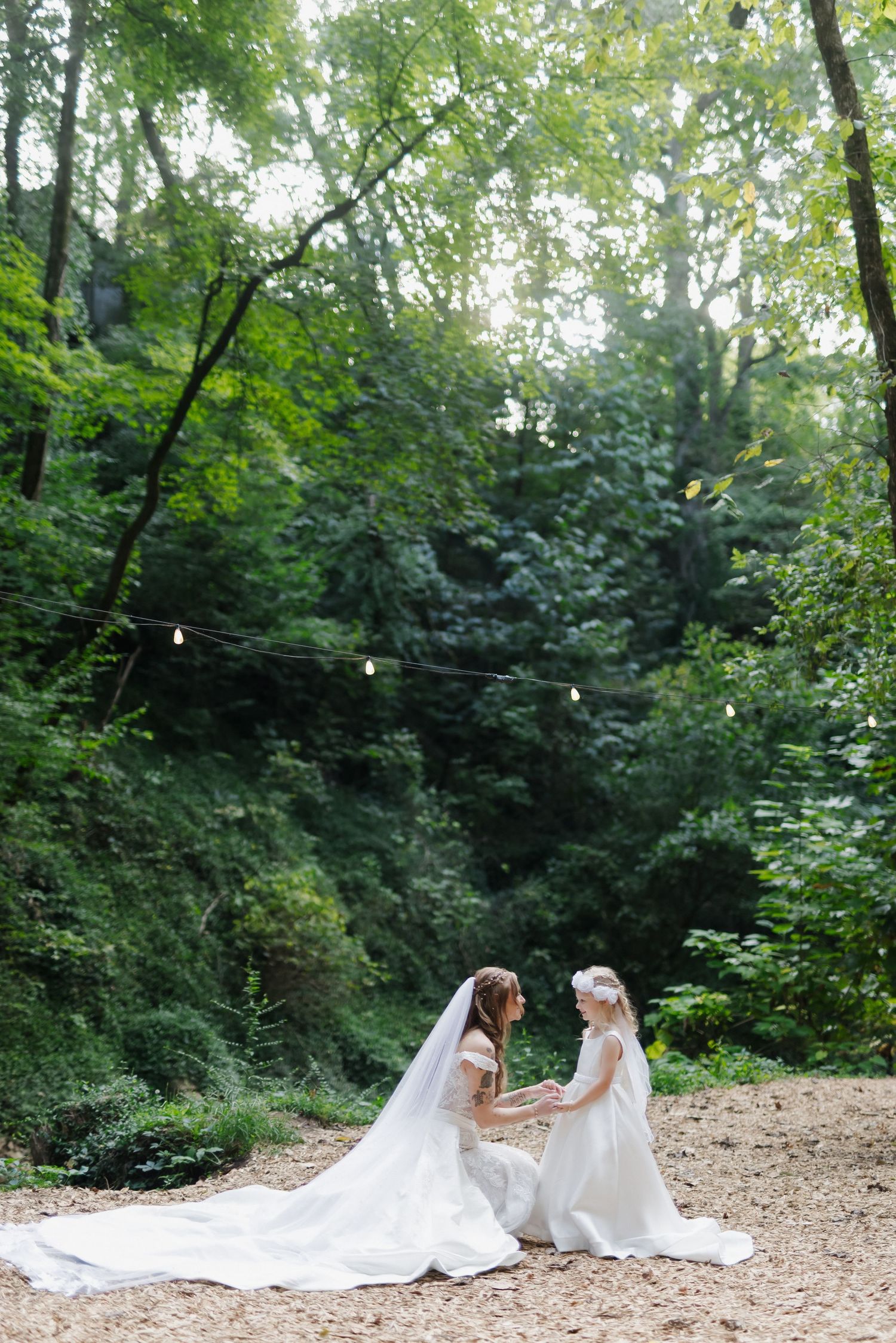 Bride in white gown with long veil kneels to hold hands with flower girl in forest wedding setting.
