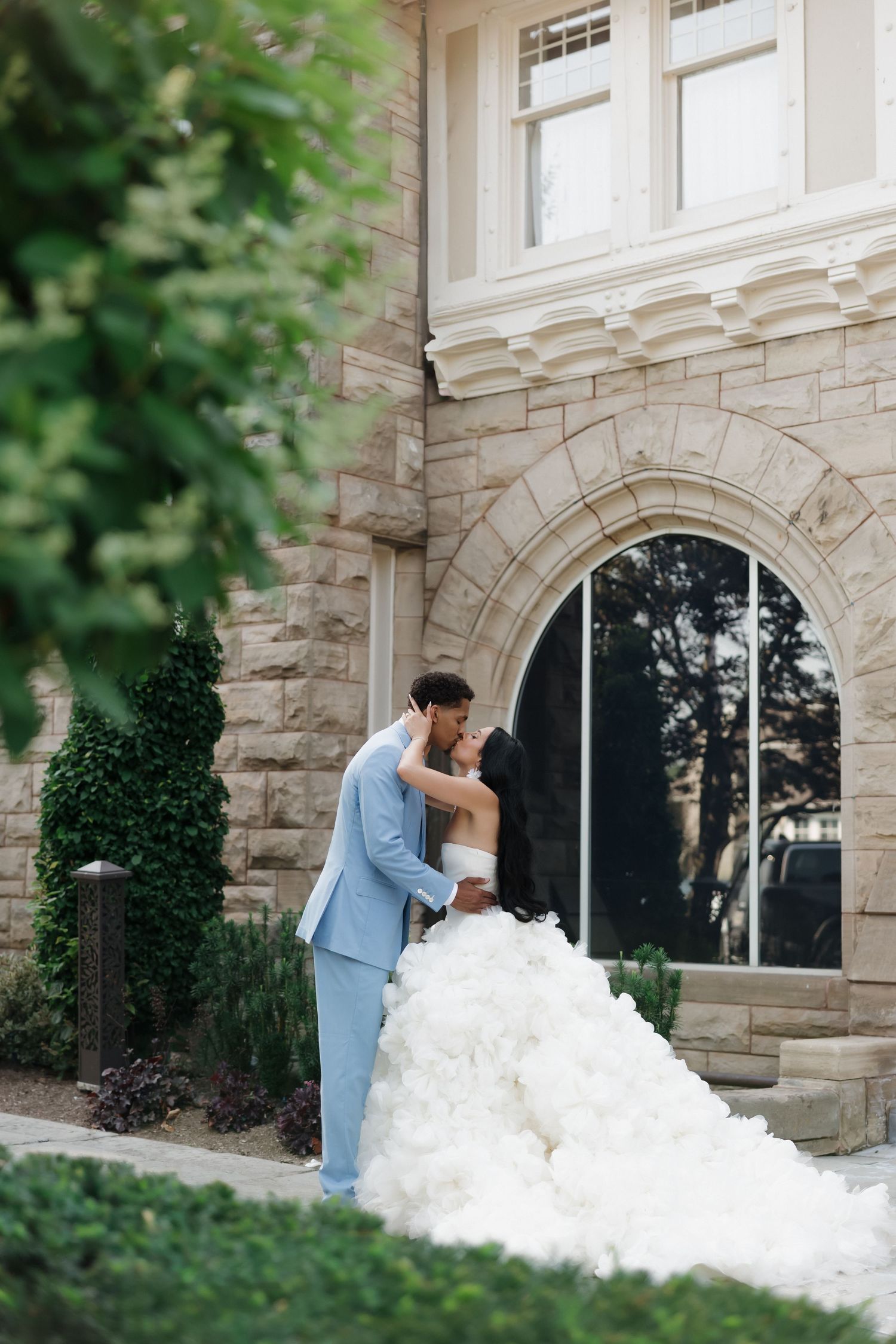 Bride in a voluminous white gown and groom in a light blue suit share a romantic kiss outside an elegant stone building.