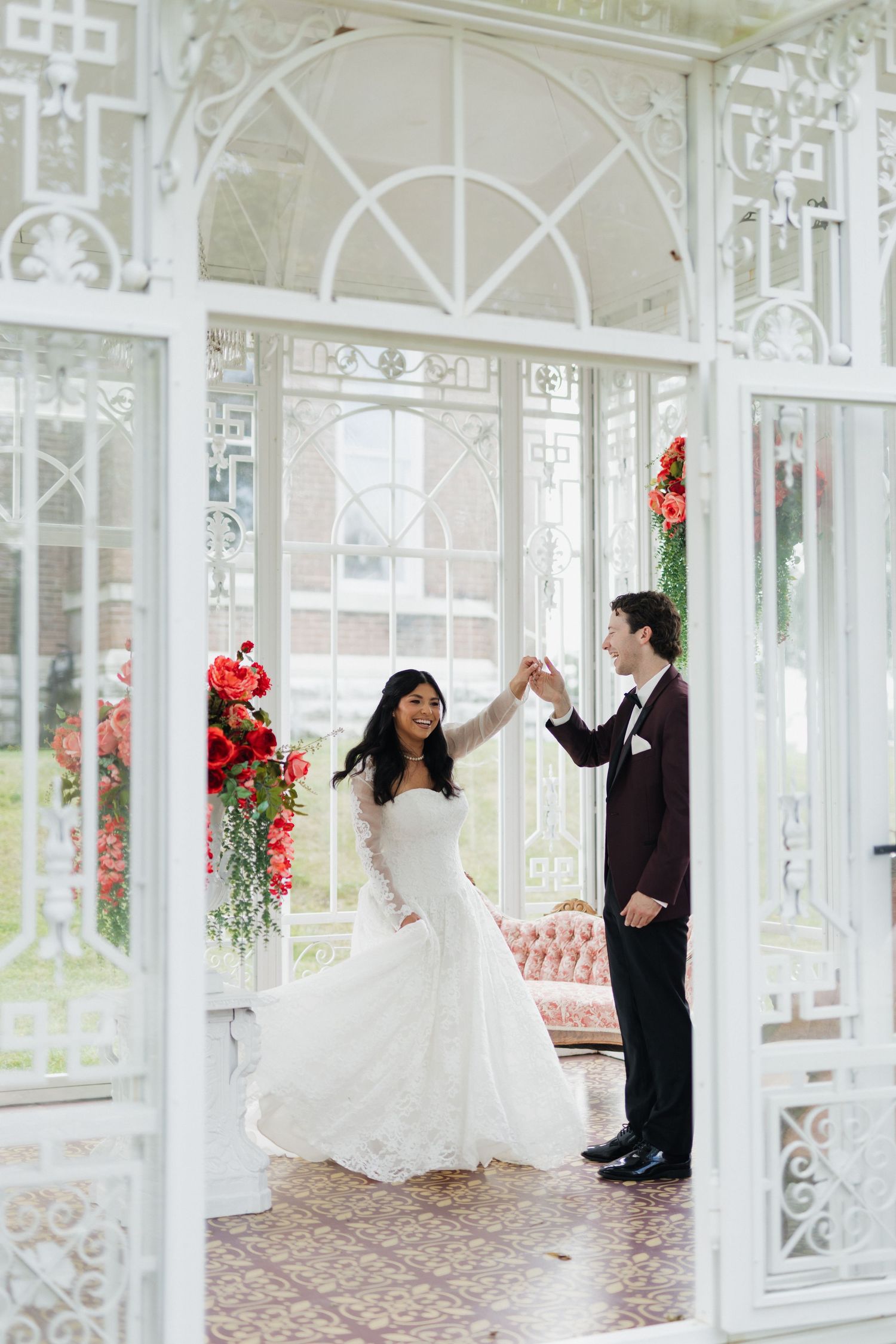 Bride and groom dancing in an elegant white Victorian greenhouse adorned with red floral arrangements.