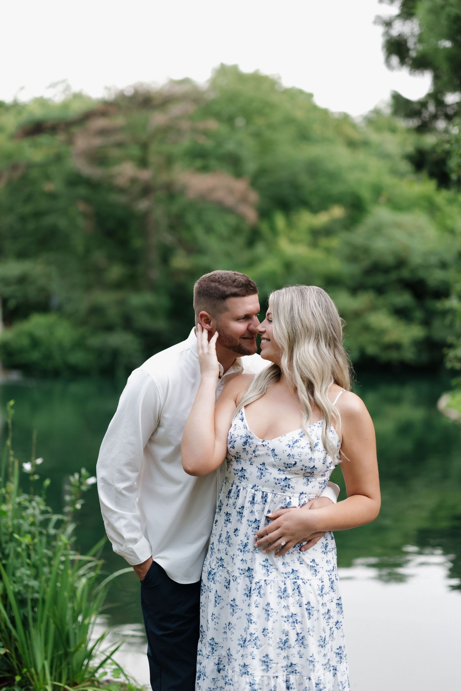 Couple sharing a romantic moment by a serene pond surrounded by lush greenery, woman in blue floral dress.