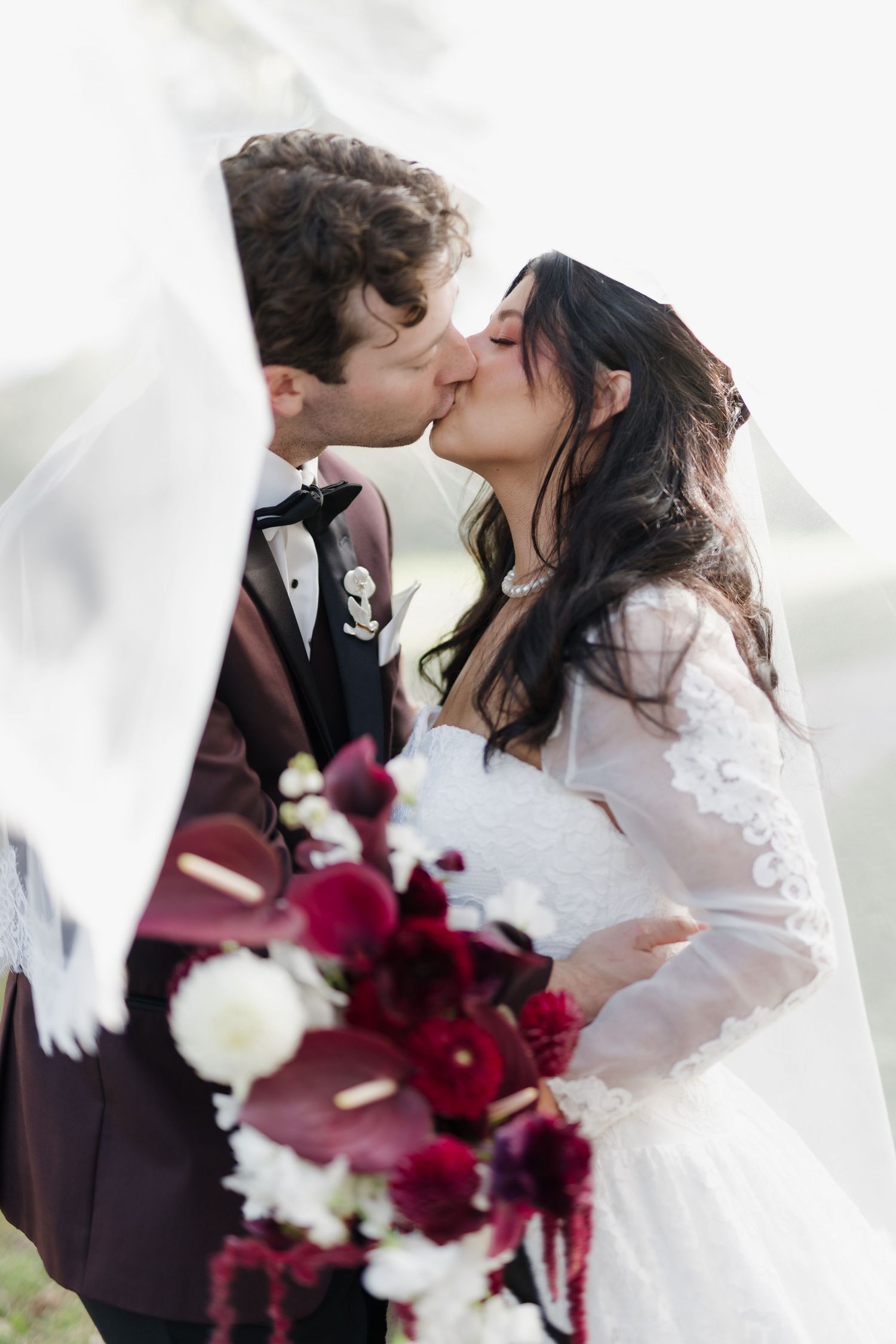 Bride and groom sharing a romantic kiss beneath a flowing veil with a burgundy bridal bouquet.