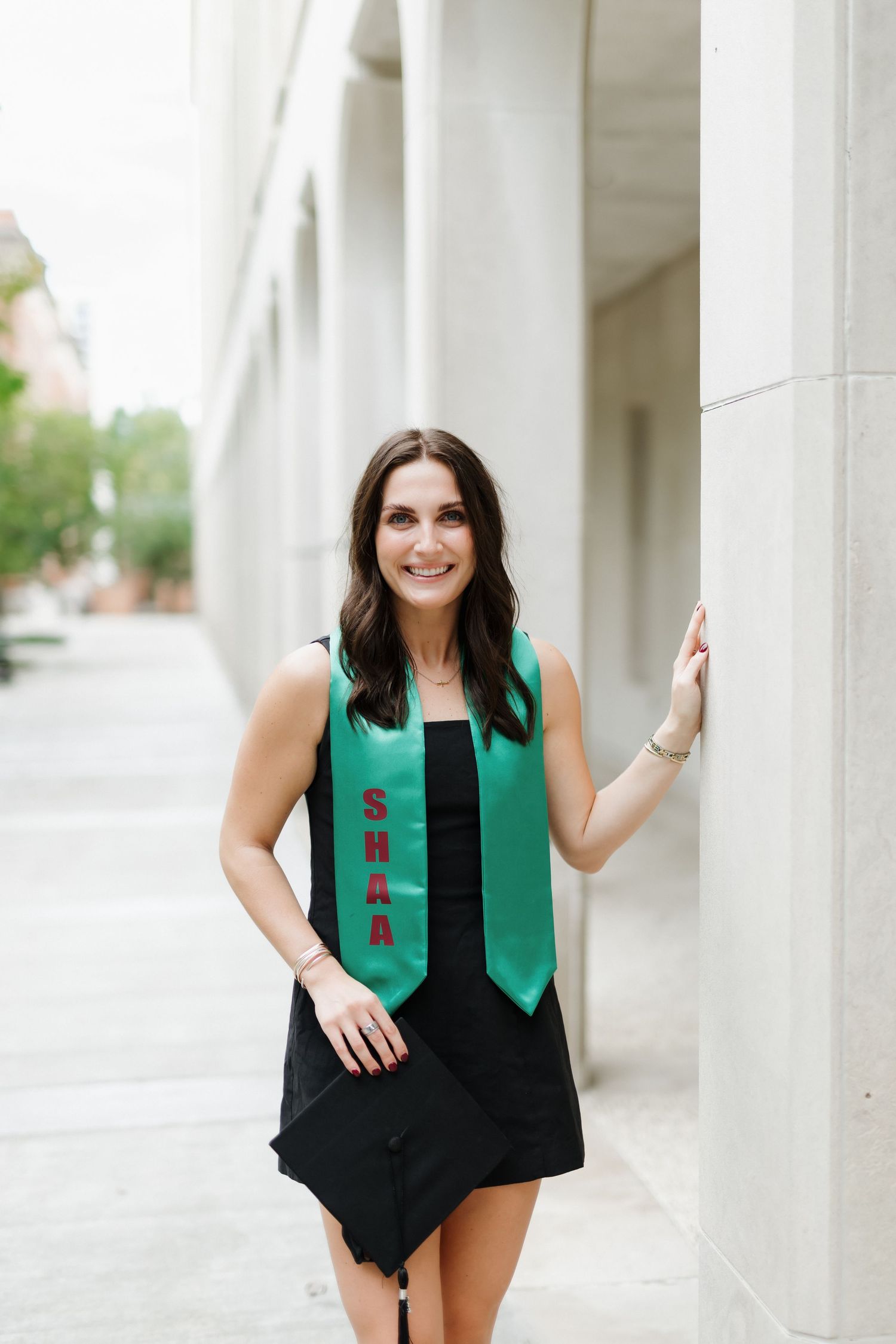 A smiling woman in a black dress wearing a green SHAA graduation stole poses outdoors near a building column.