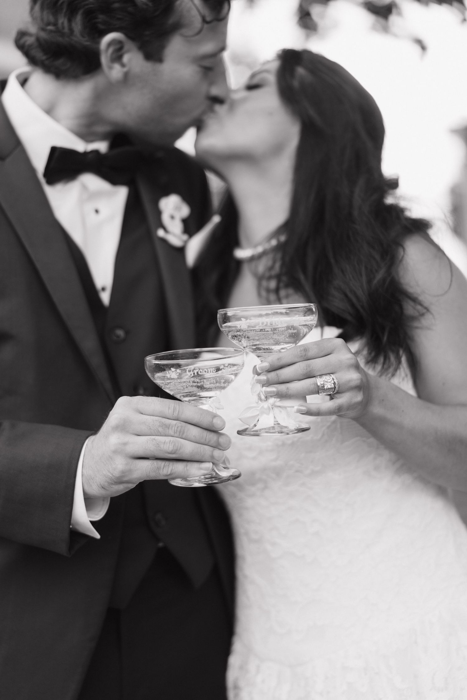 Black and white photo of a bride and groom kissing while holding champagne glasses at their wedding reception.