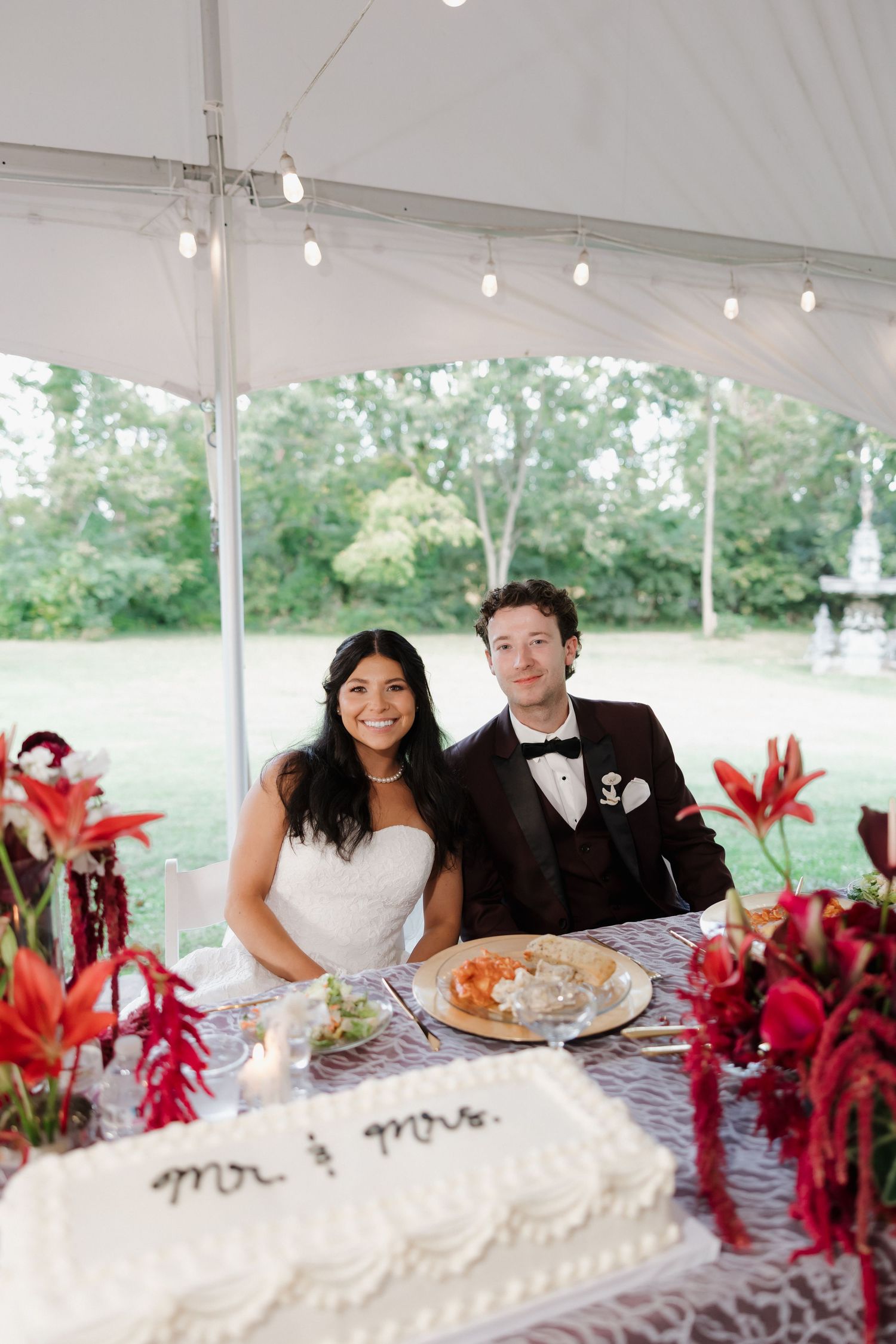 Bride and groom smile at sweetheart table with white Mr. & Mrs. cake and red floral arrangements under tent.