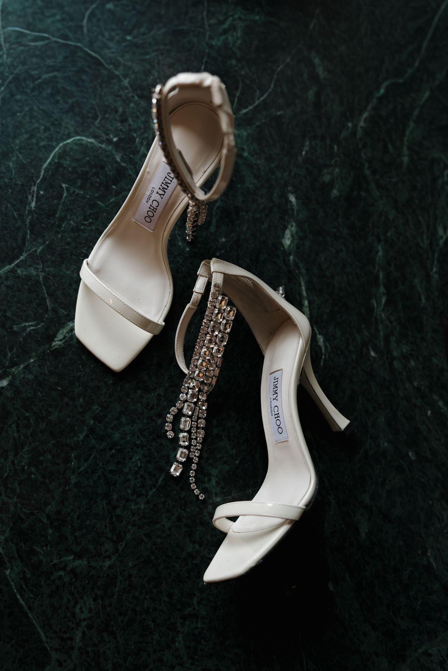 Elegant Jimmy Choo white bridal heels with crystal embellishments on dark green marble surface.